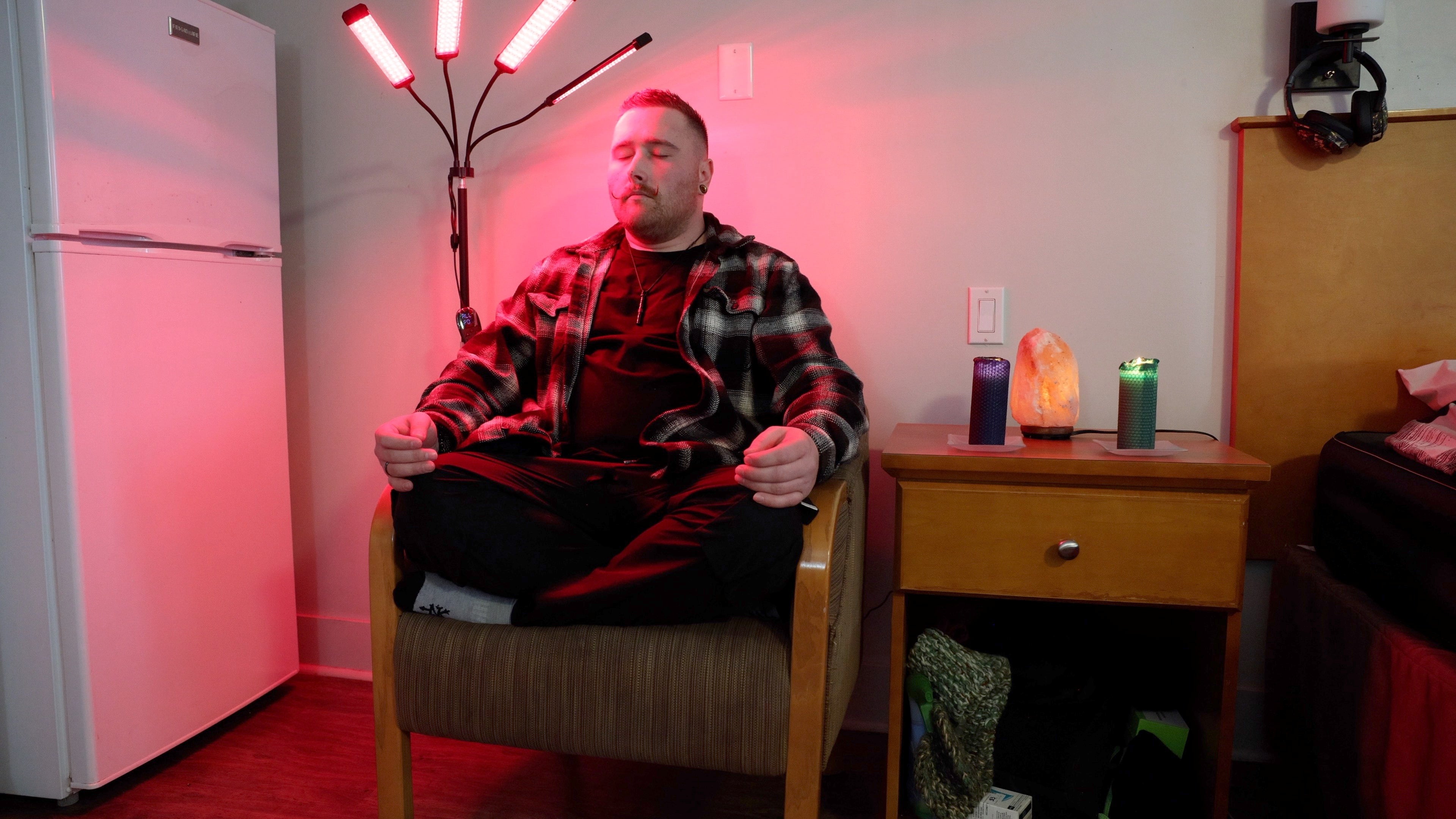 A person with short brown hair and a curled moustache sits cross-legged in a chair meditating underneath infrared light.