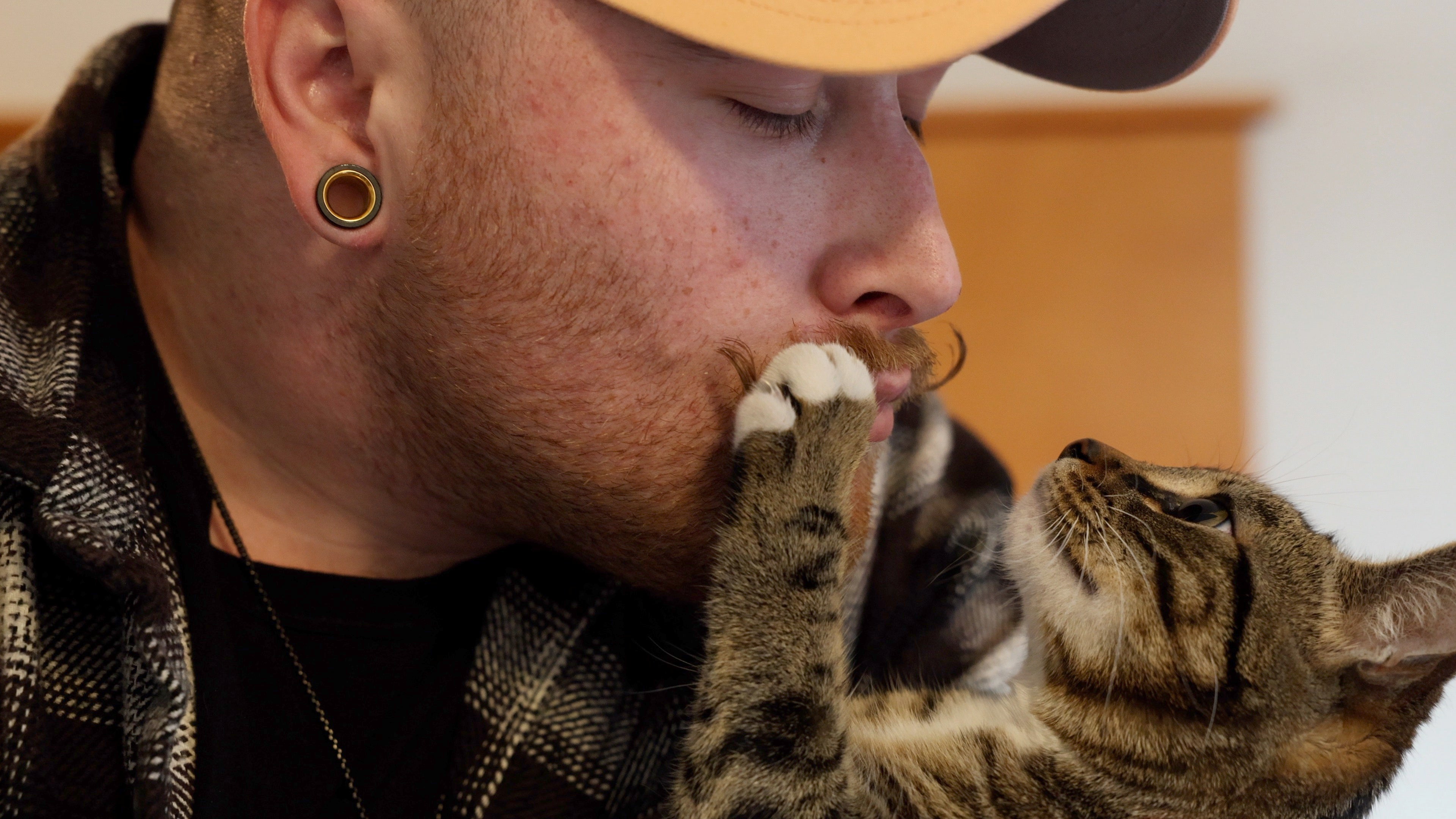 A person cuddles and looks lovingly into a cat’s eyes as it grabs their moustache.