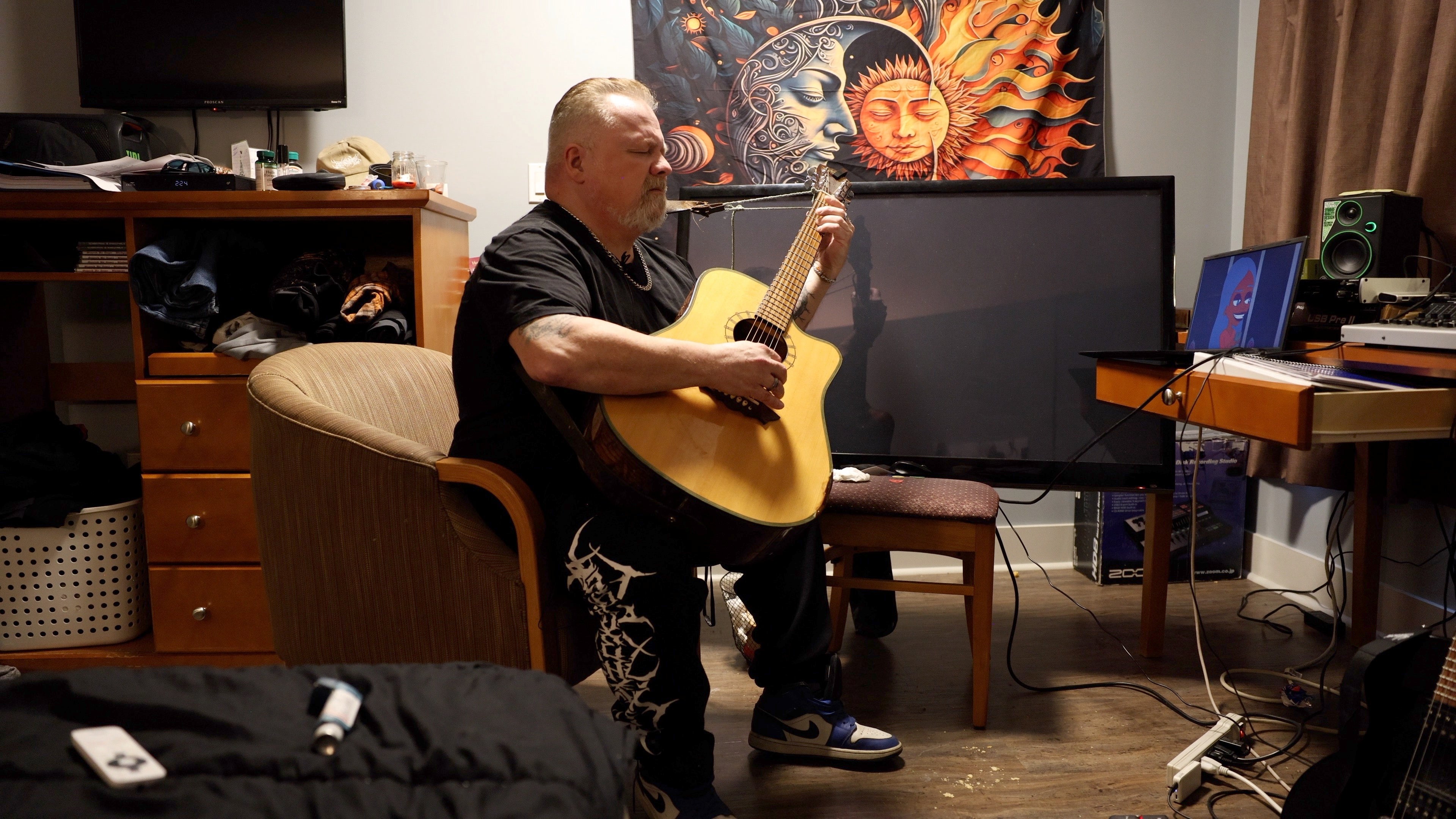 A person with a goatee in their room playing an acoustic guitar and singing.