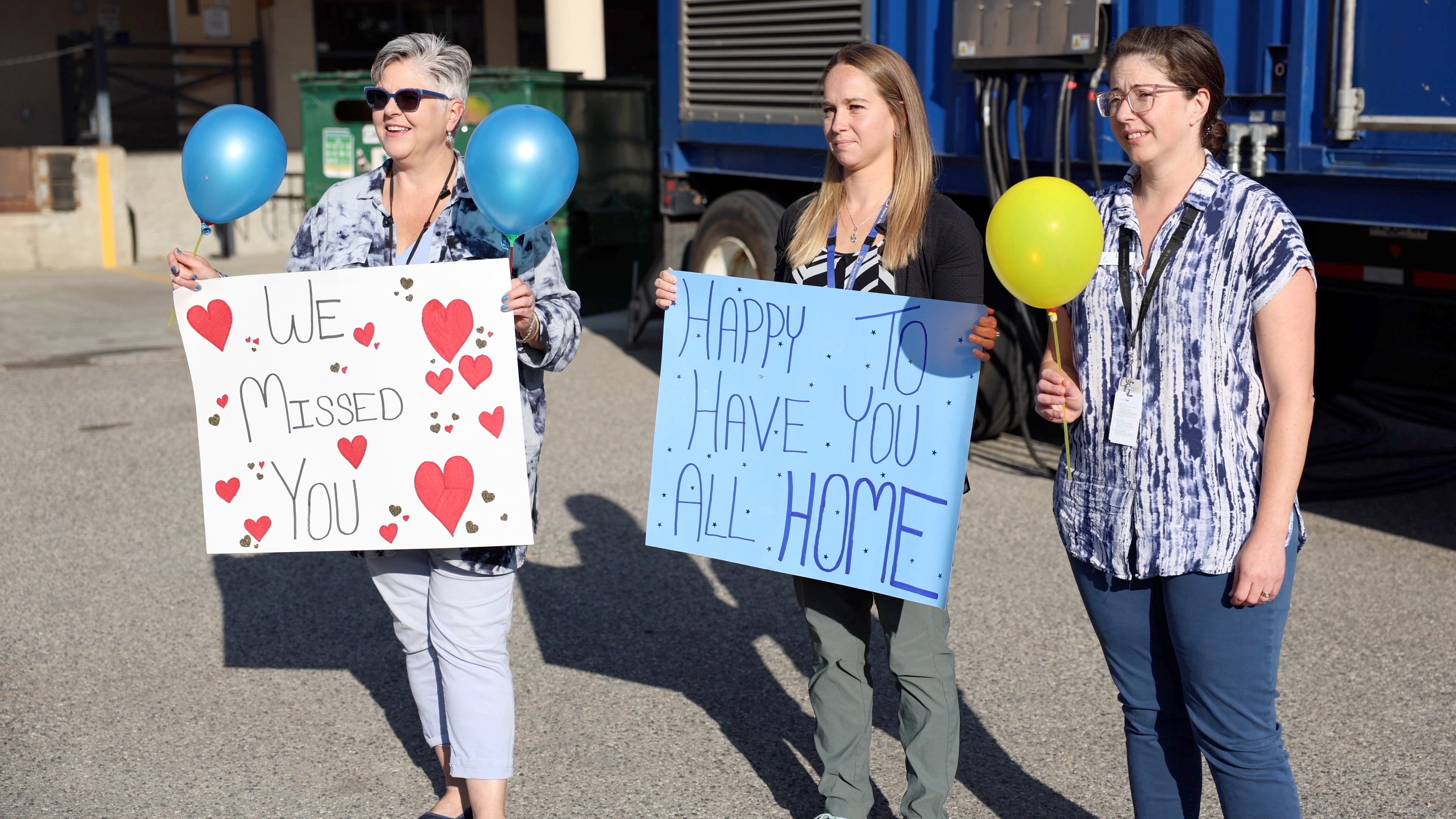 Three people stand outside holding balloons and two signs, one with hearts that says we missed you, and another with stars that says happy to have you all home.