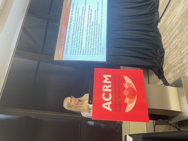 A woman with long blond hair wearing glasses and a white t-shirt stands in front of a podium that says ACRM next to a screen showing a presentation