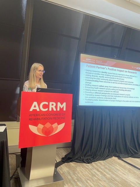 A woman with long blond hair wearing glasses and a white t-shirt stands in front of a podium that says ACRM next to a screen showing a presentation