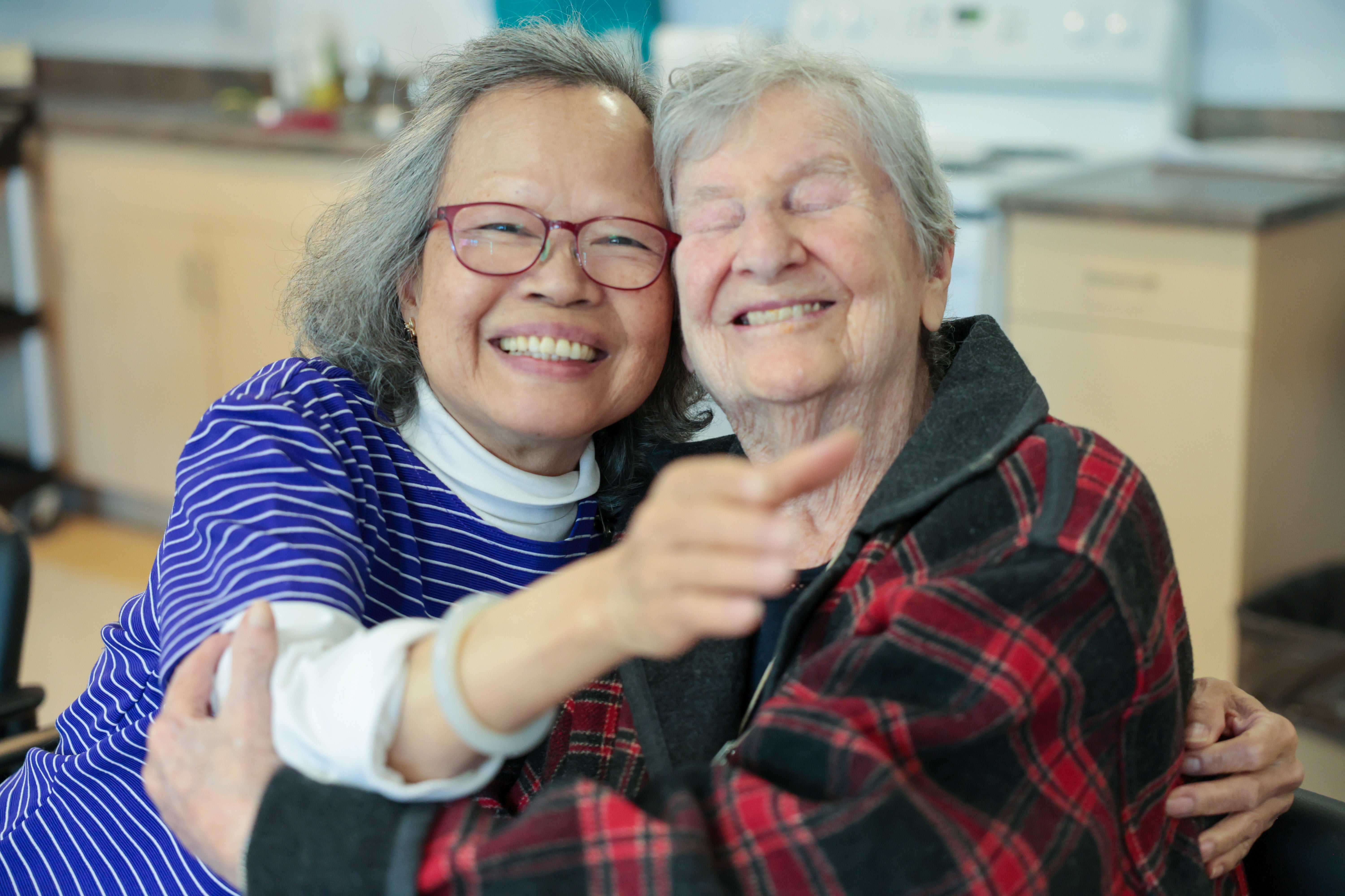 Two smiling women, one with grey hair and glasses in a purple striped shirt and the other with grey hair and eyes closed in a red plaid shirt, embrace each other