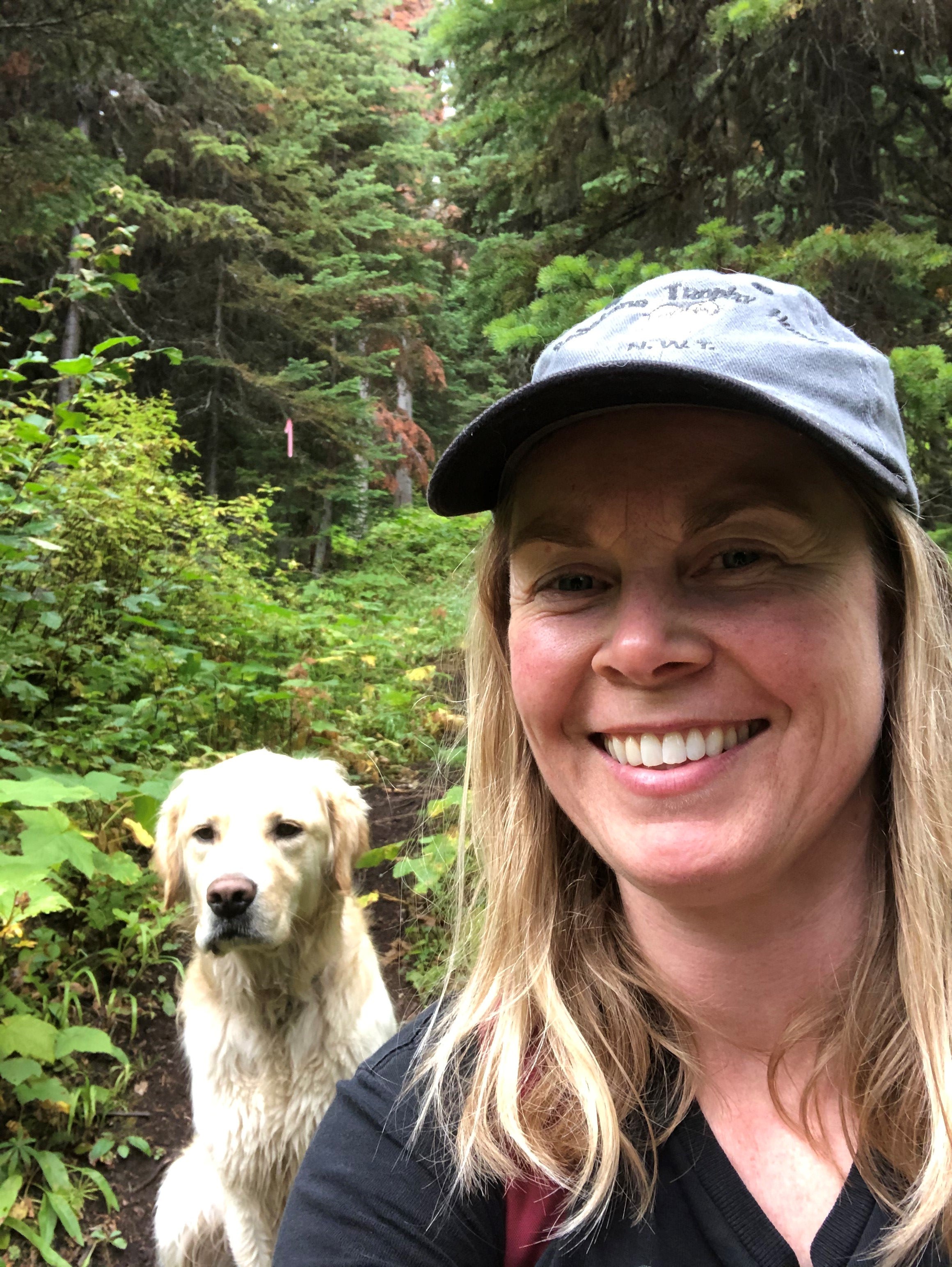 A smiling woman with long blond hair in a blue ball cap takes a selfie in a forest with her cream coloured dog behind her.