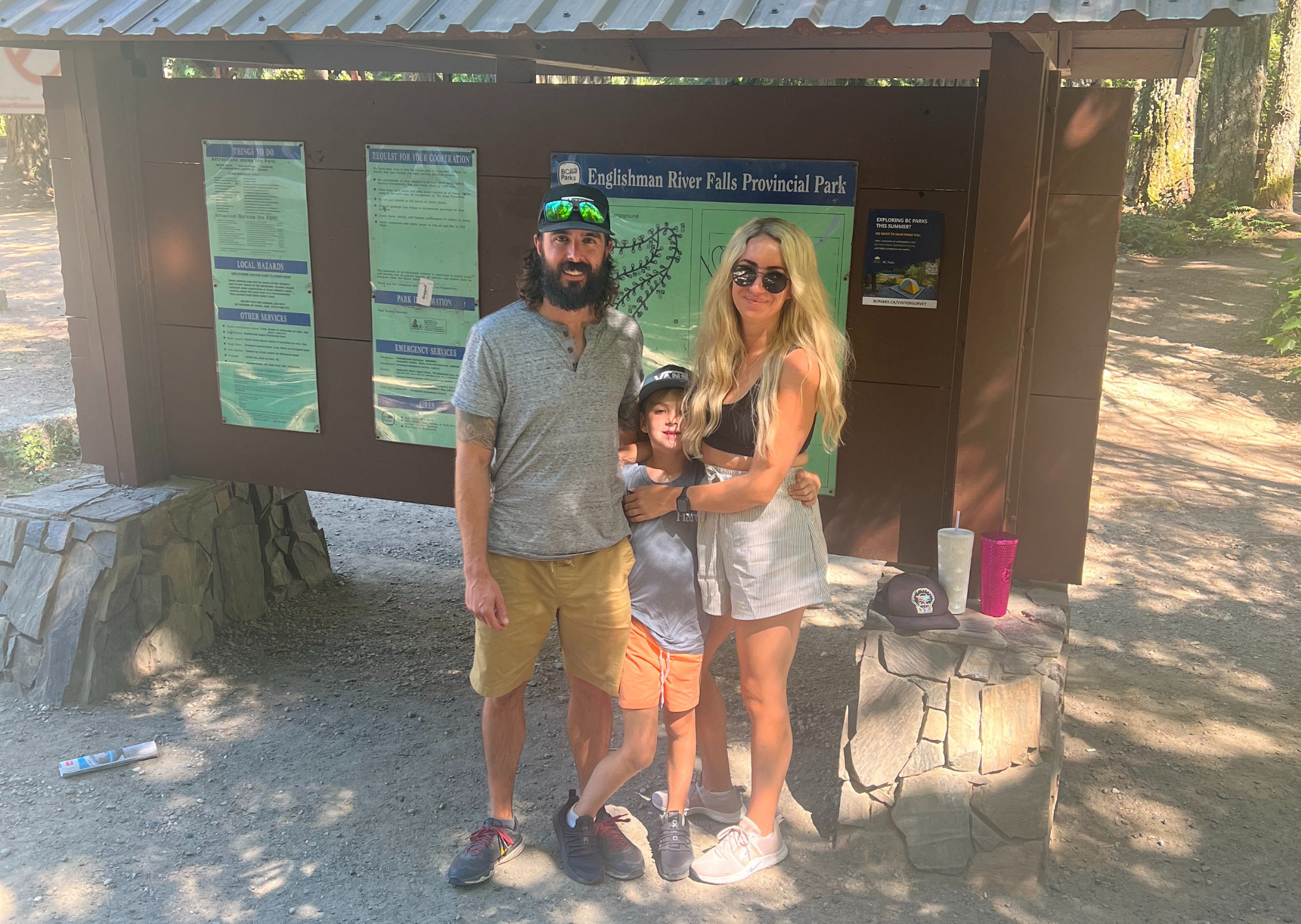 A blonde woman stands with her brunette husband and young son by a hiking route map, outdoors.