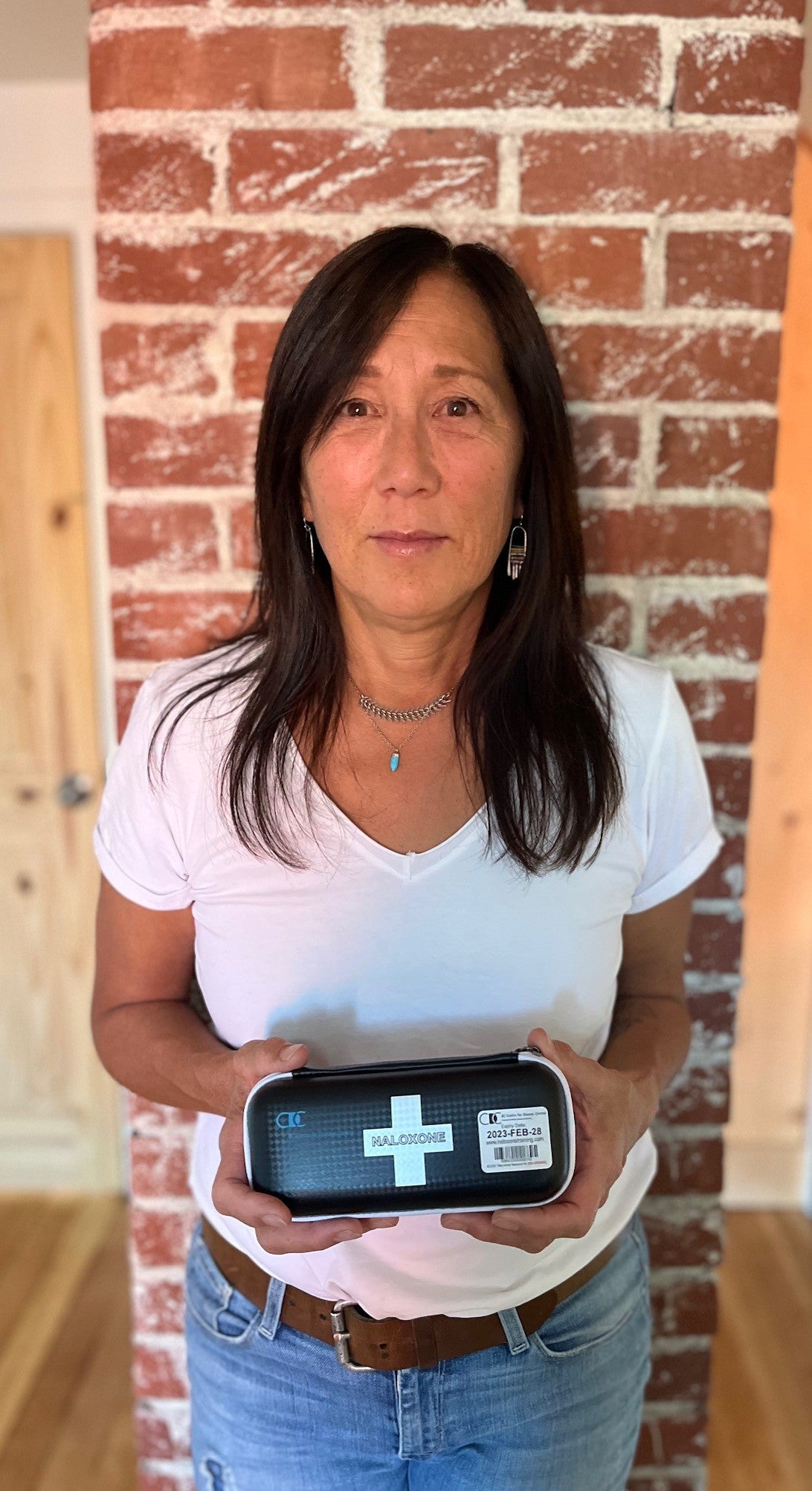 A woman with long black hair wearing a white v-neck t-shirt, jeans and silver jewellery stands against a brick wall holding a black, oval naloxone kit