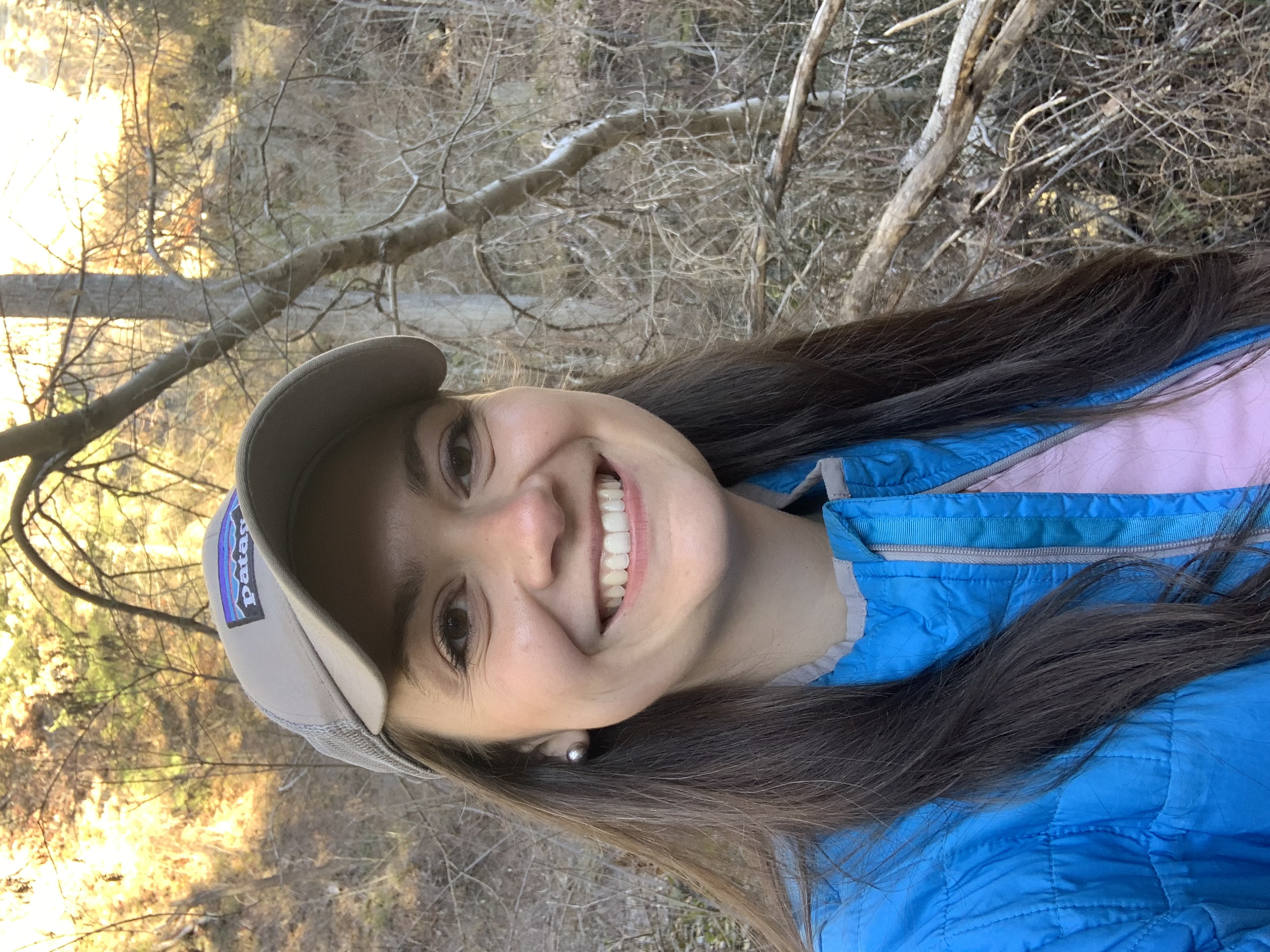 A smiling woman with long dark hair wearing a tan ballcap and blue jacket takes a selfie in a forest