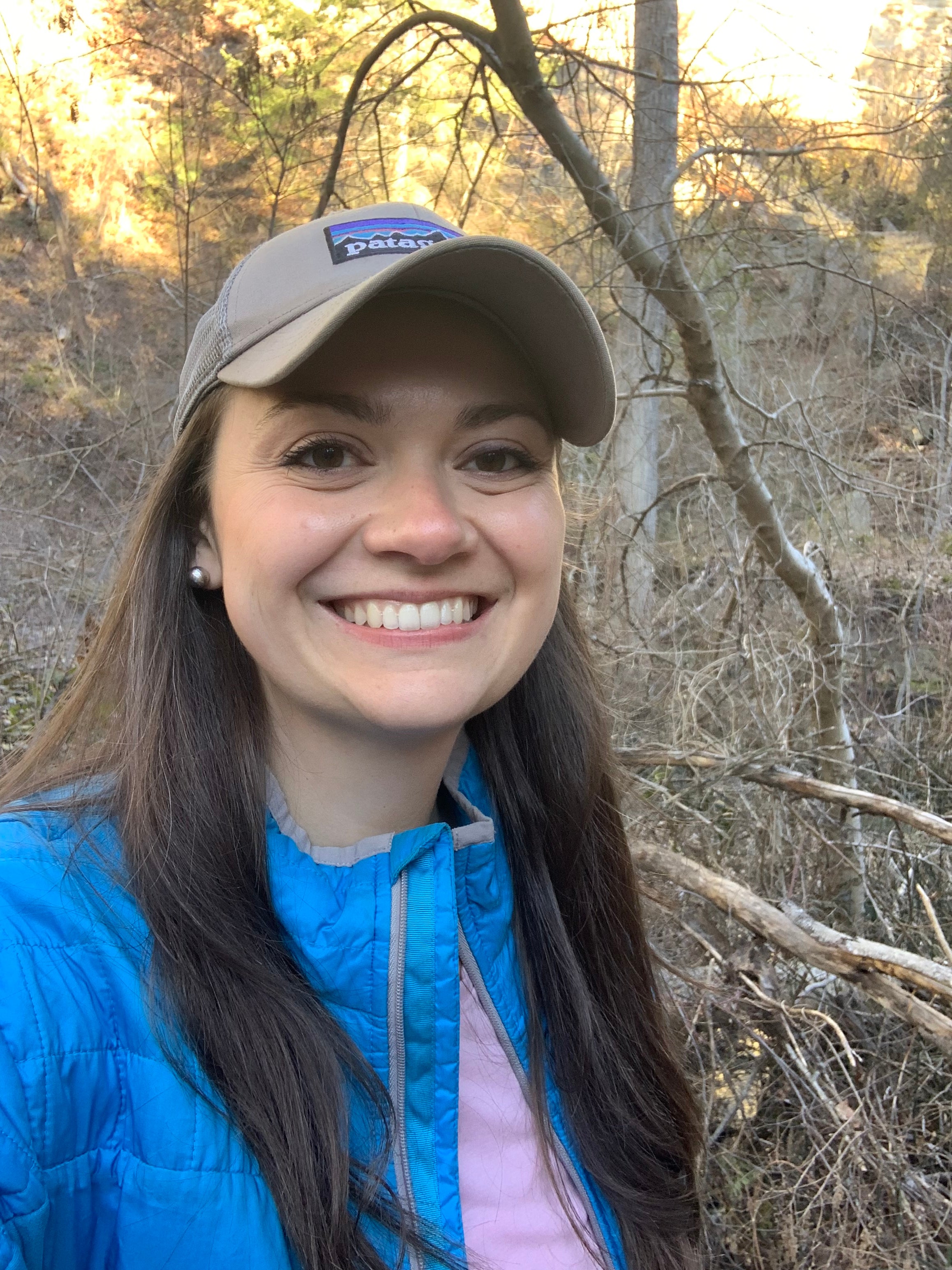 A smiling woman with long dark hair wearing a tan ballcap and blue jacket takes a selfie in a forest