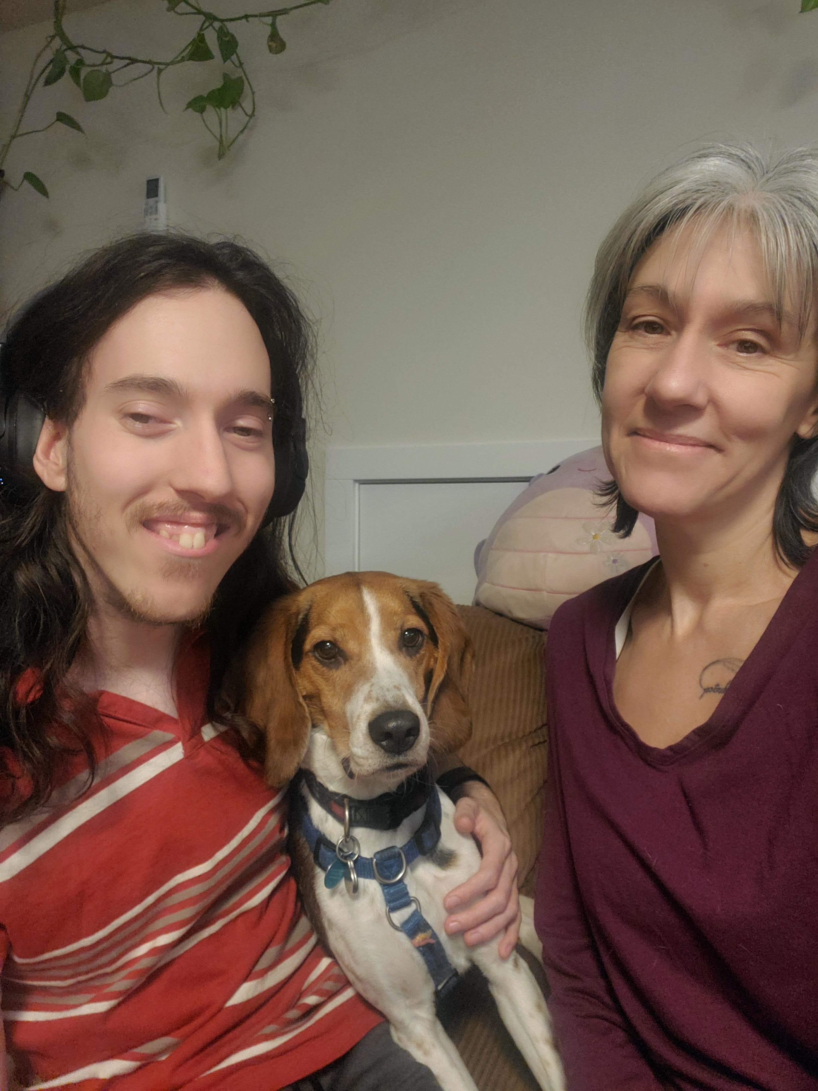 A young man with long brown hair wearing a red striped shirt holds a brown and white beagle next to a smiling woman with grey medium-length hair wearing a burgundy top