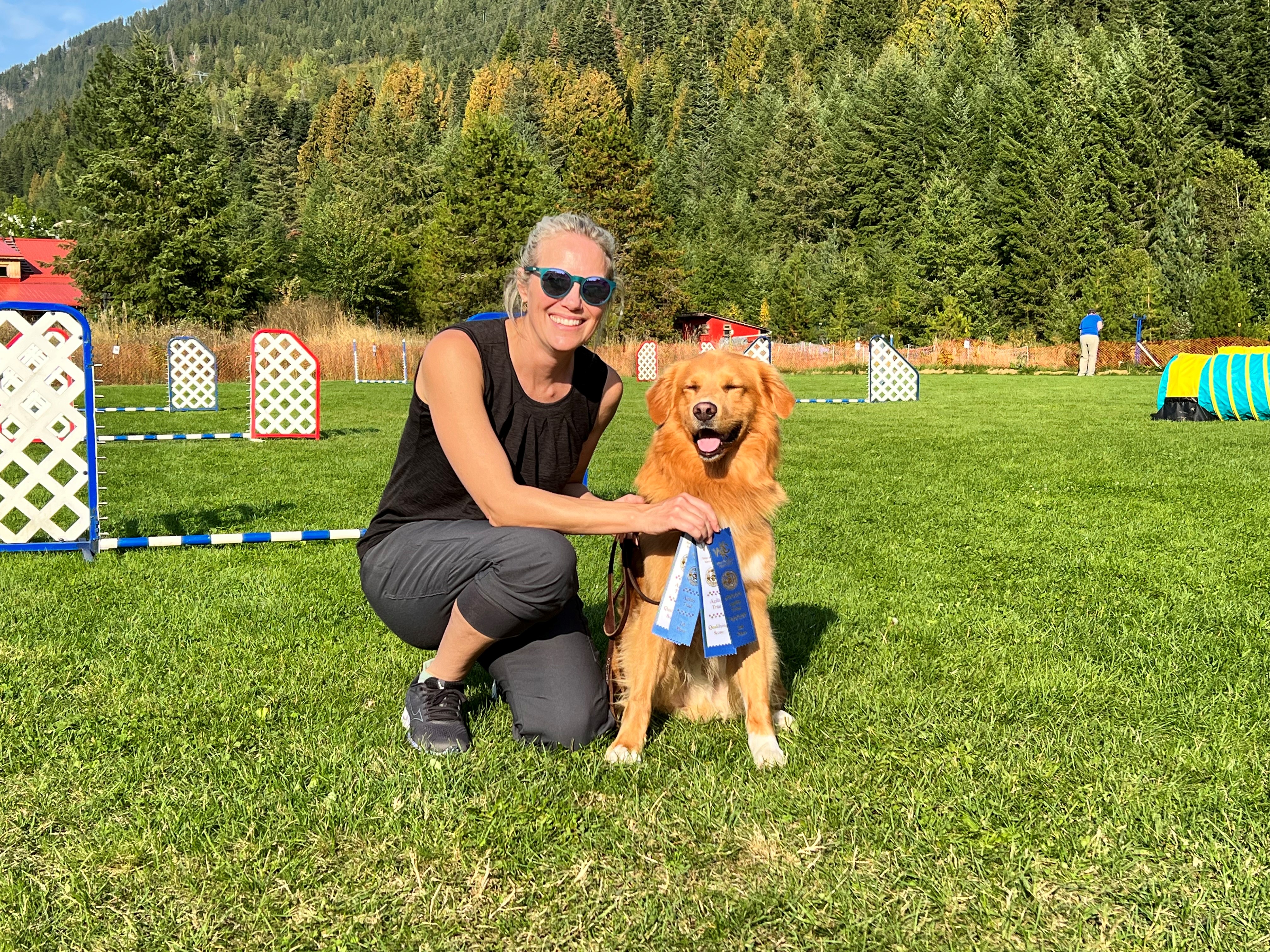 A smiling woman in a black tank top and grey pants holds a blue ribbon in front of an orange dog on a grassy field