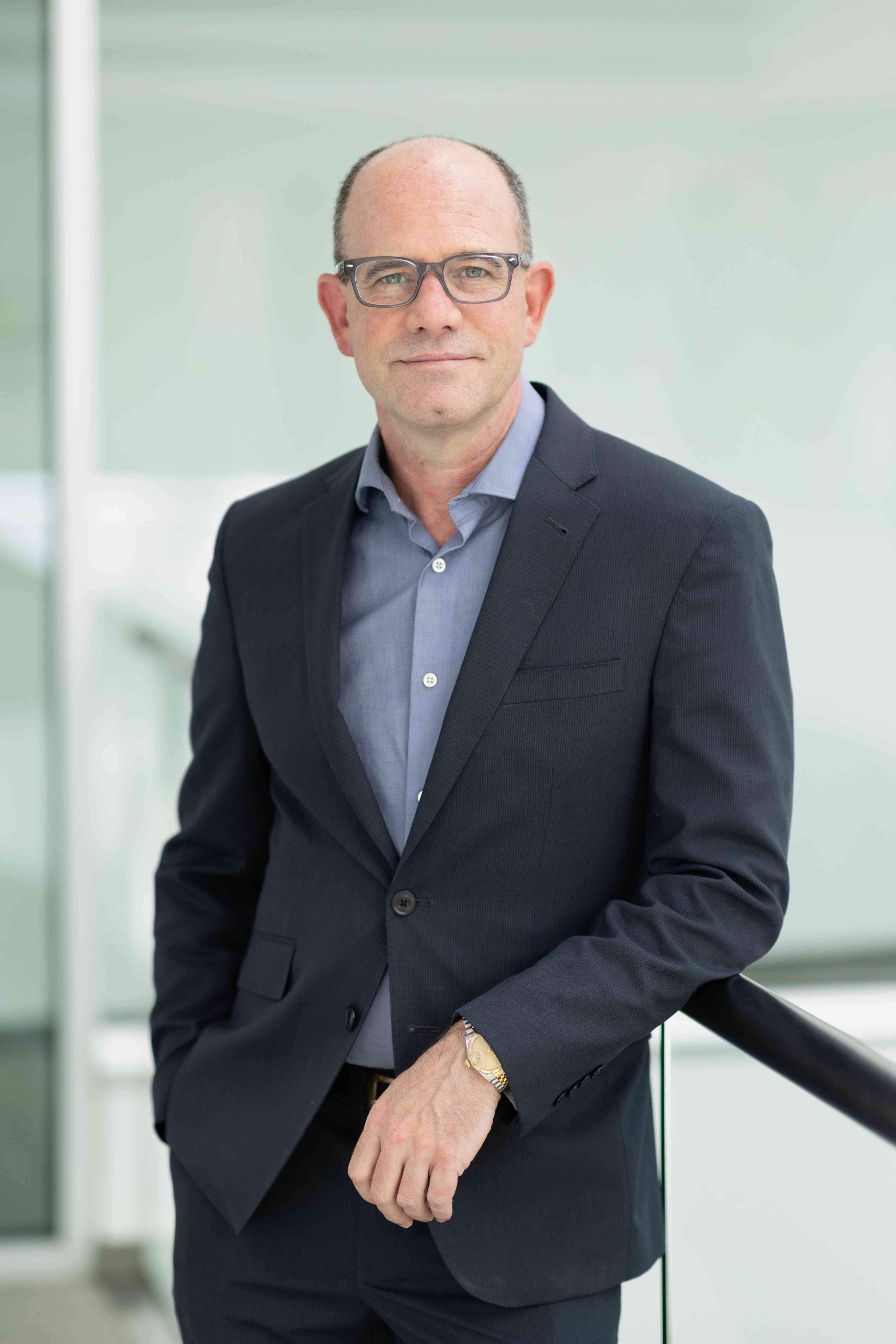 A business profile picture of a person with glasses wearing a suit jacket.