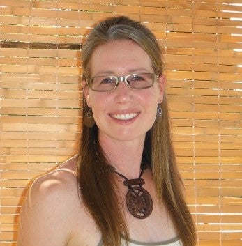 A smiling person with long hair that's tied back, and wearing silver eyeglasses, a tank top and a necklace with a large wooden charm.