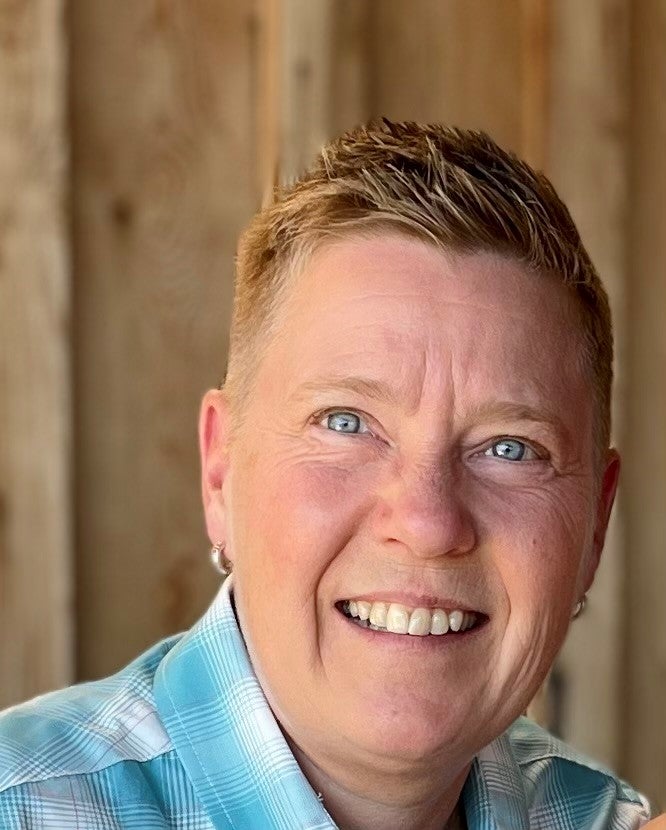 A profile picture of a smiling person with short blonde hair and blue eyes, who is wearing a light blue plaid shirt and posing in front of a wooden wall background.