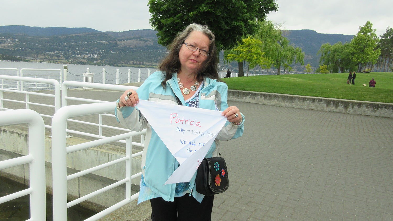 A woman standing on a sidewalk near water holds a white flag that says "Patricia" in red letters.