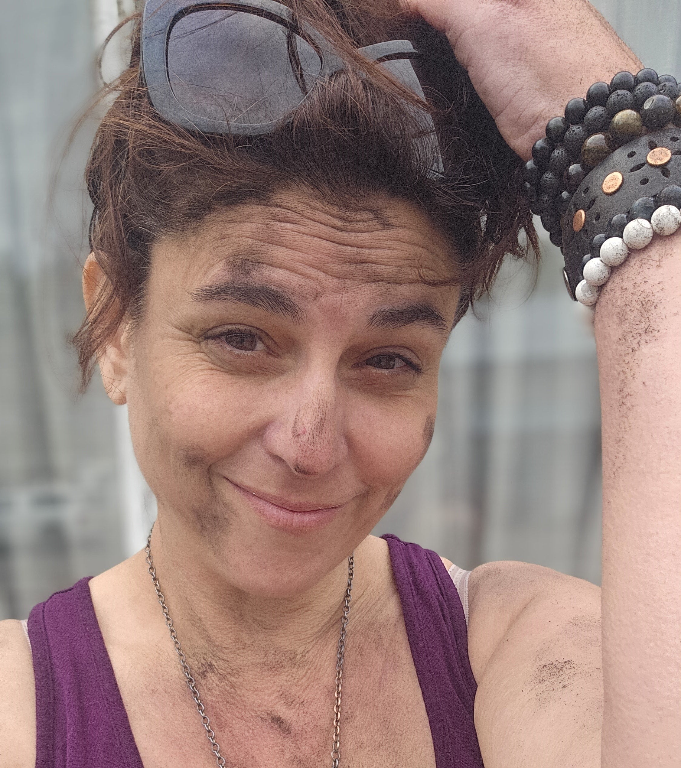 A smiling person with a dusty face rests their arm on their knee. They’re wearing a burgundy tank top, bangles and sunglasses on their head.