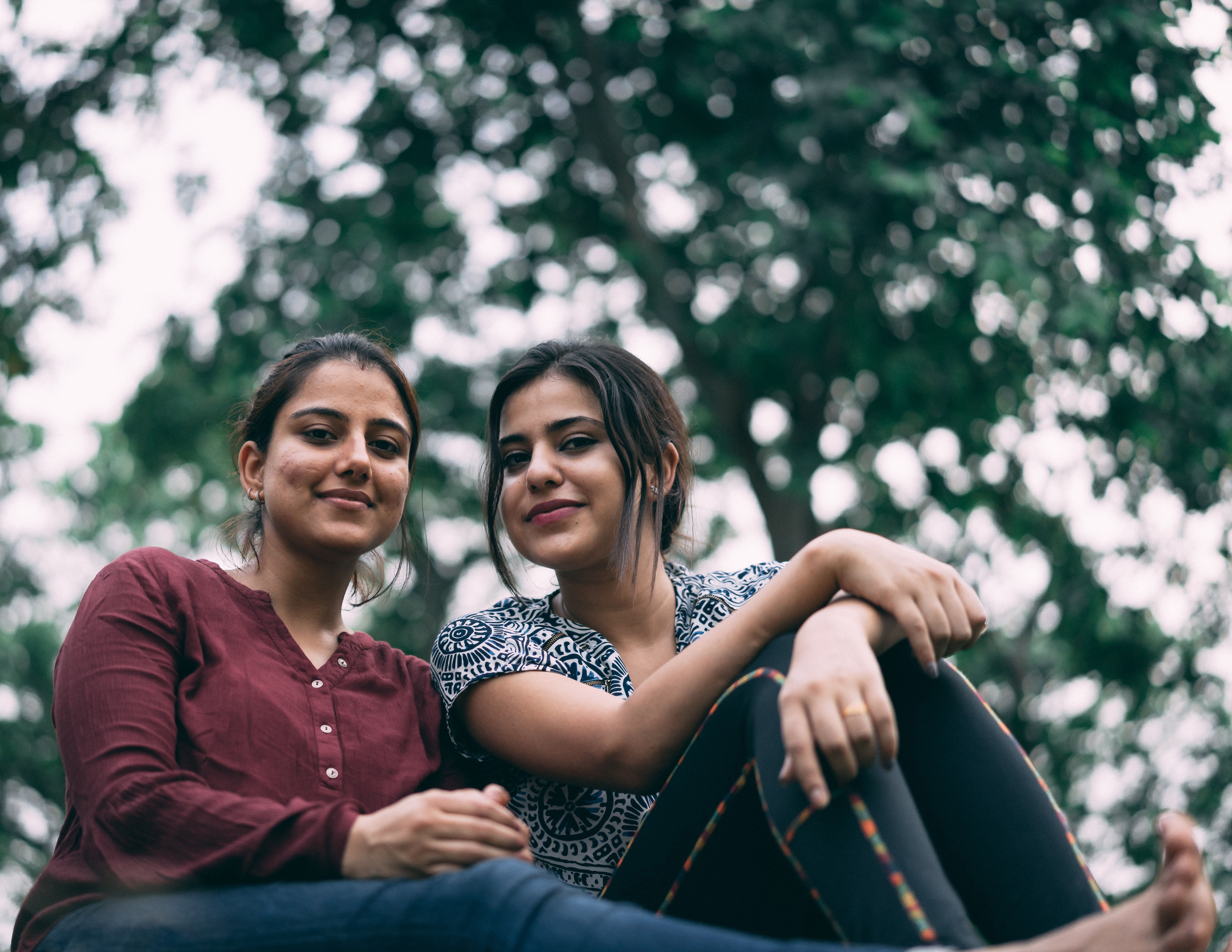 Two young people sit casually together. The photo is taken outside with blurred trees in the background.