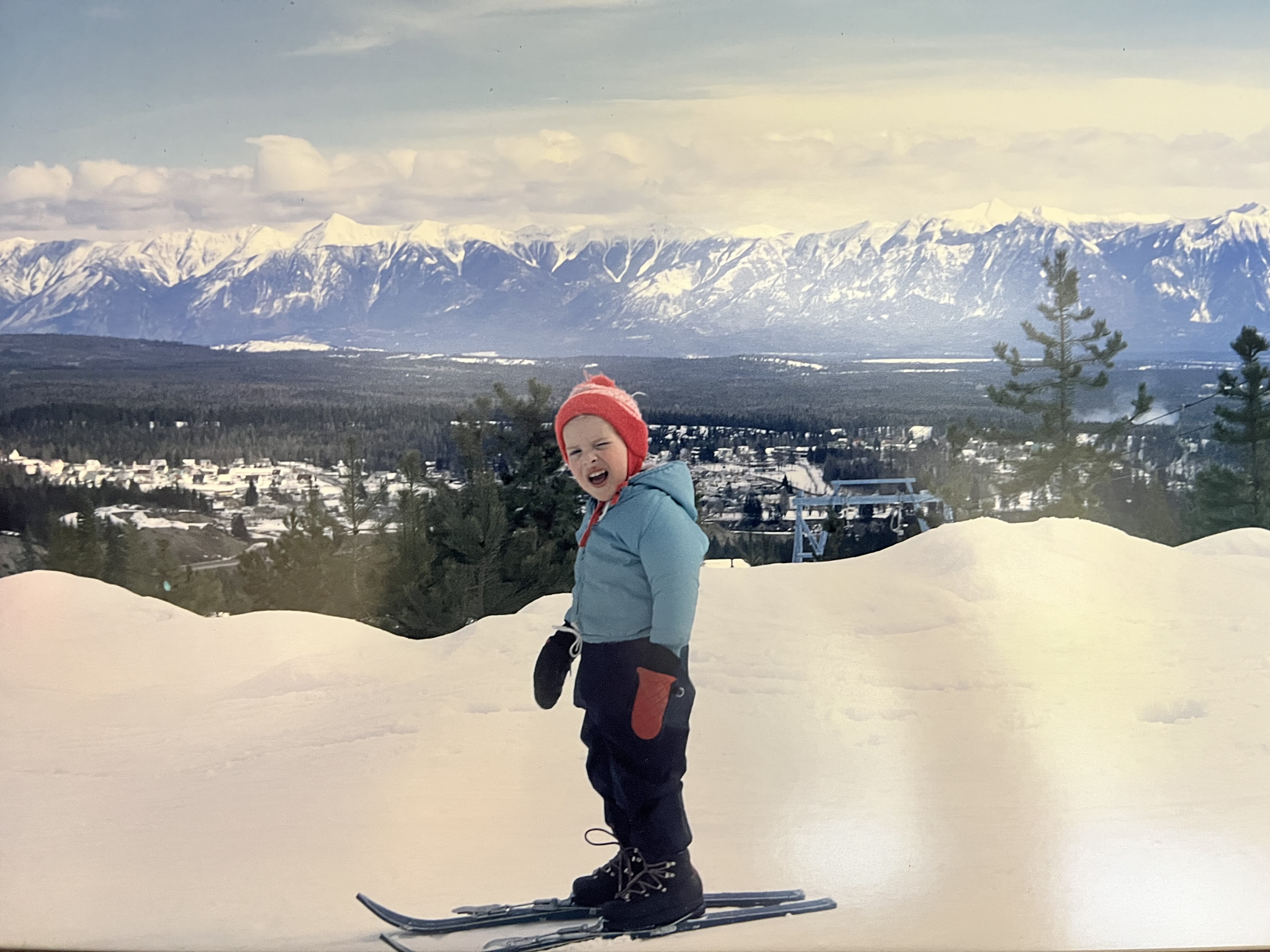 A young child in a red toque and mittens, and blue ski jacket, stands on skis on a hill with a mountain range in the background