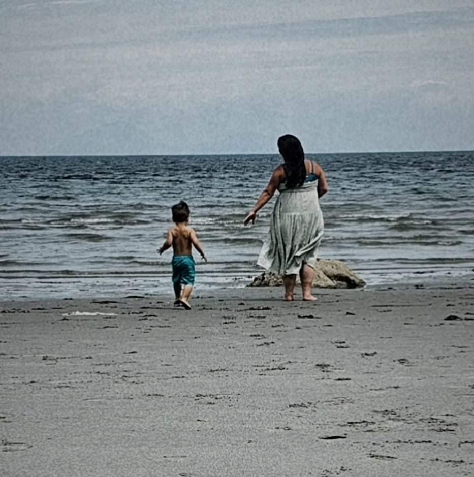 A woman in a black swimsuit top and white skirt runs beside a boy in blue swim trunks on a beach