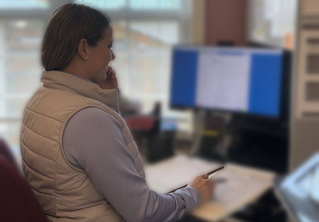 A person in a white vest and light blue long-sleeved shirt sits at a desk with a monitor.