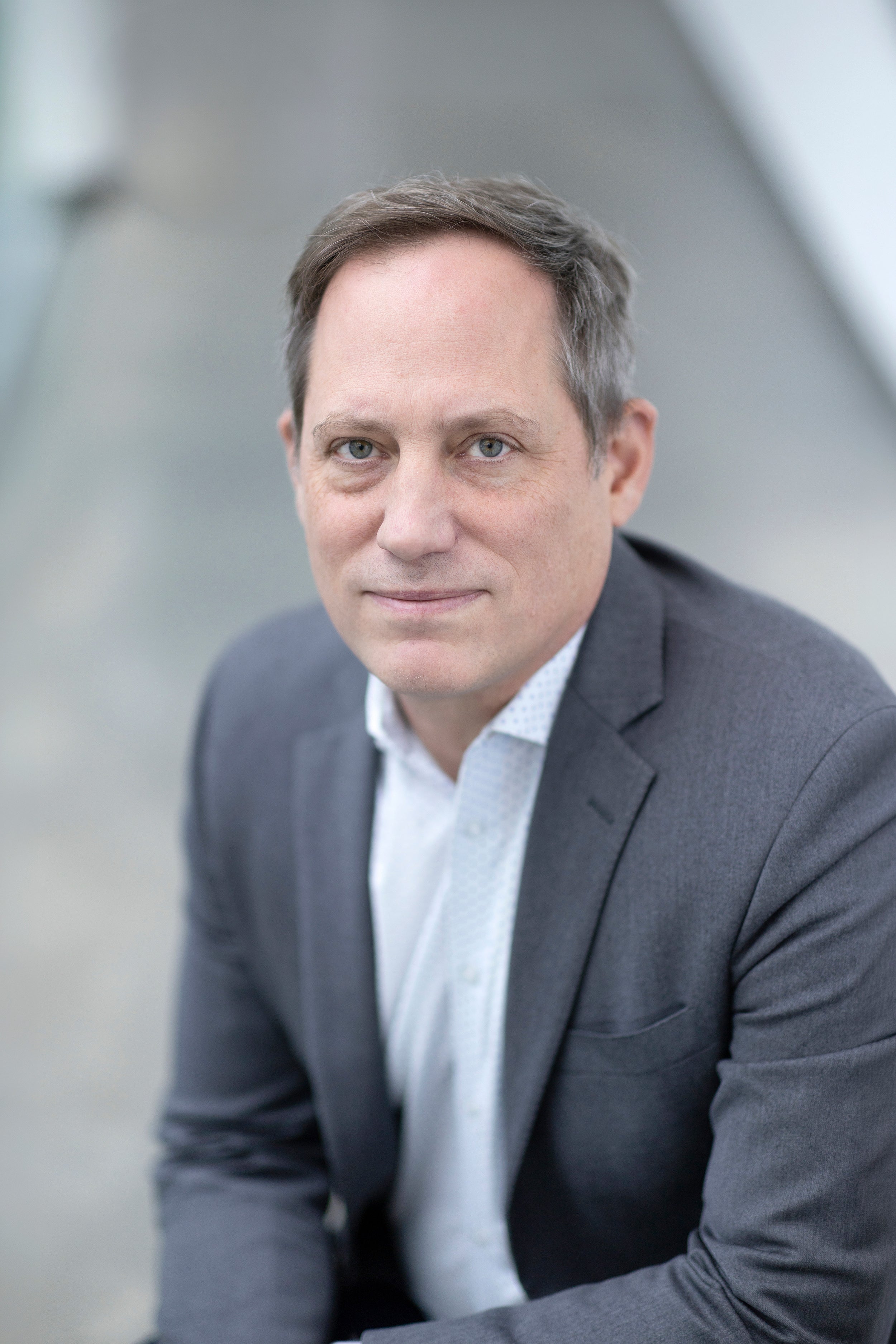 Profile shot of a person with short hair wearing a light blue shirt and grey suit jacket.