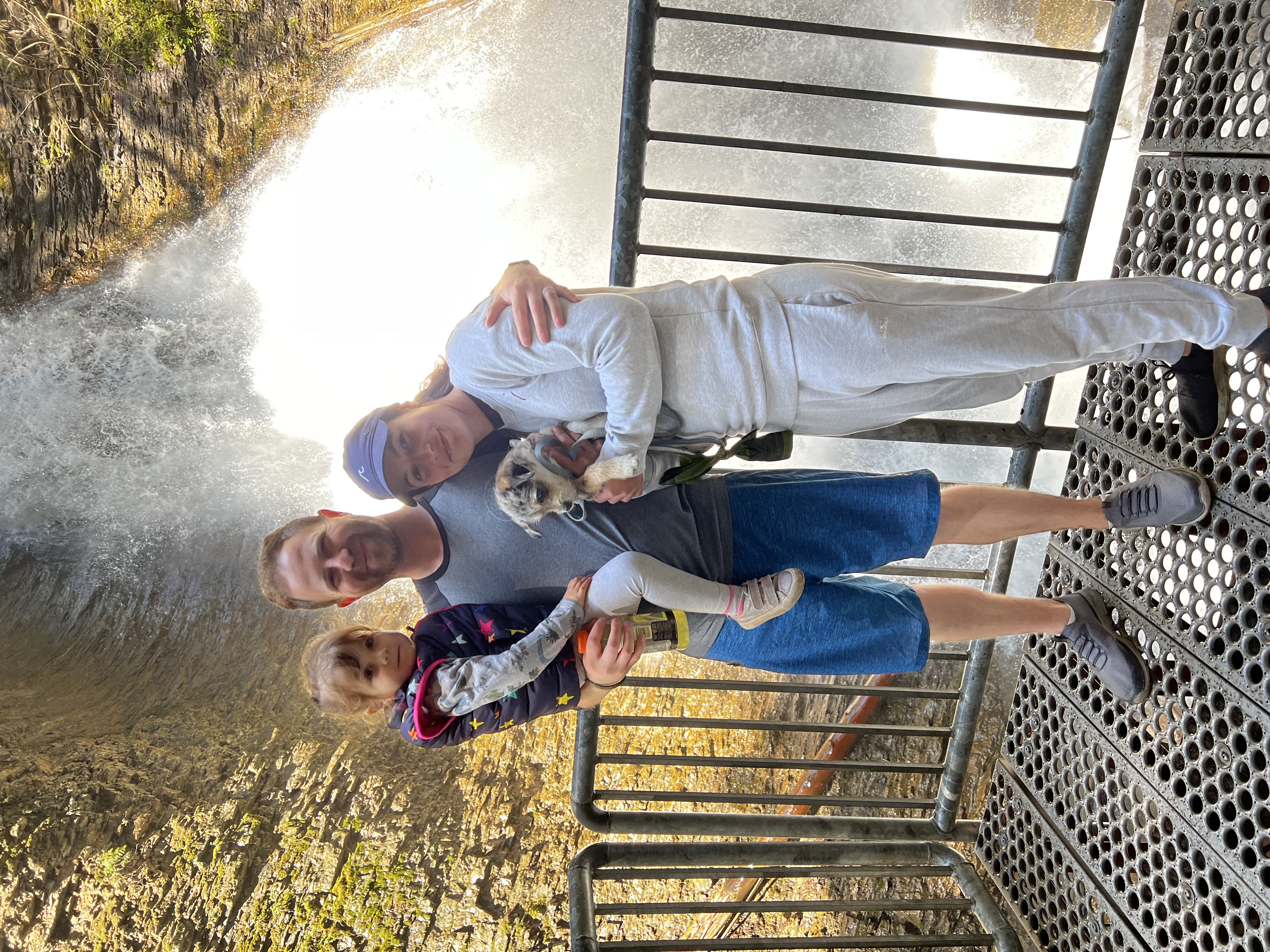 A mom in white shirt and pants, a dad in blue shorts and tshirt and a child in his arms stand together on a metal platform in front of a waterfall