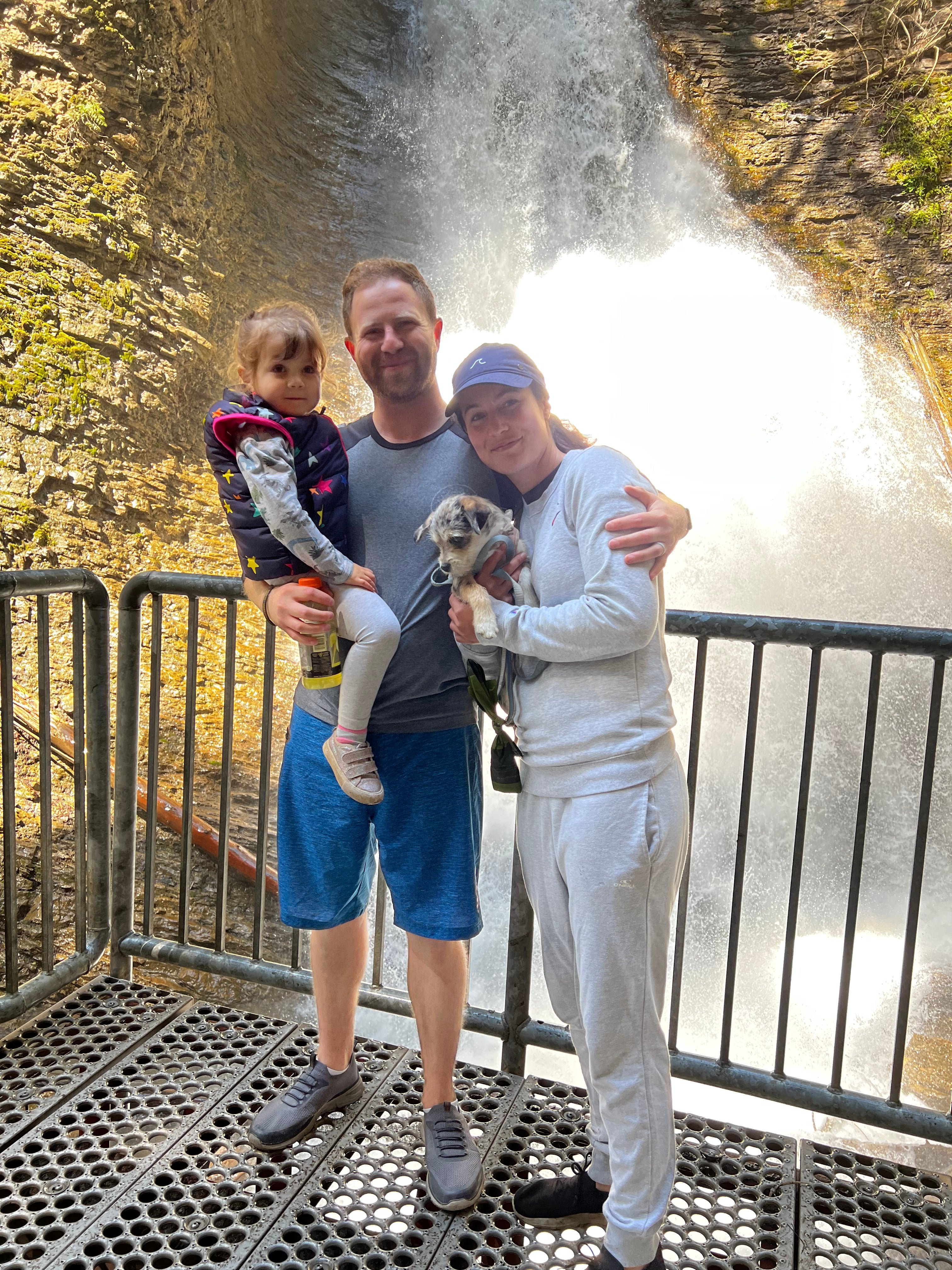 A mom in white shirt and pants, a dad in blue shorts and tshirt and a child in his arms stand together on a metal platform in front of a waterfall