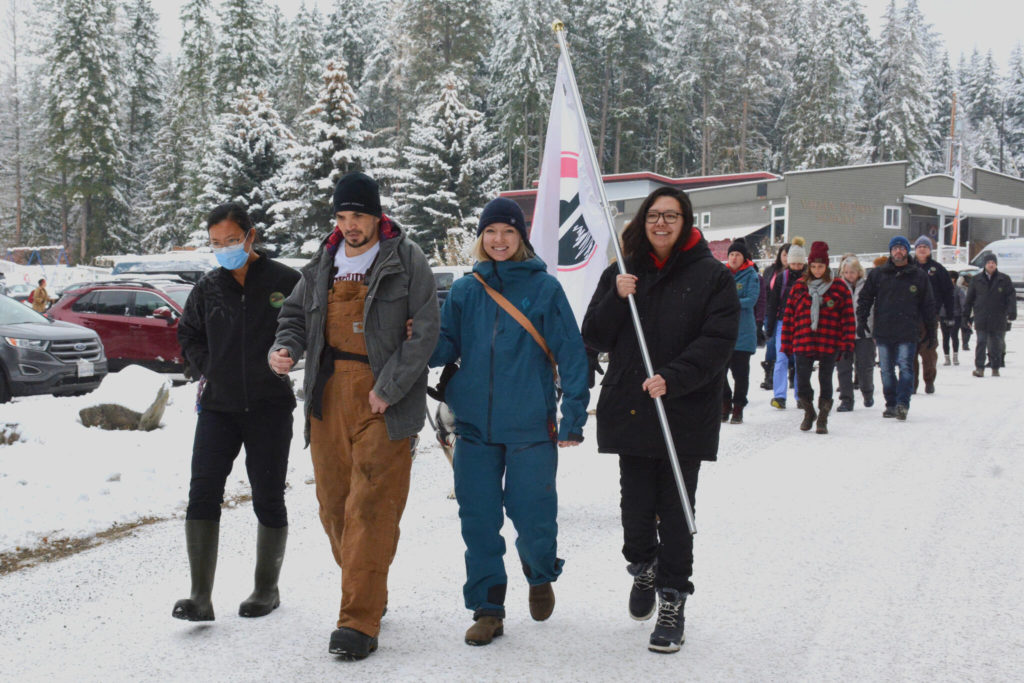 Four people wearing winter clothes walk on a snowy road. One holds a white flag.