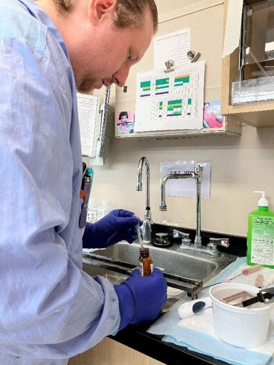 A person in a blue gown and blue gloves leans over a sink as they hold a dropper and brown vial.