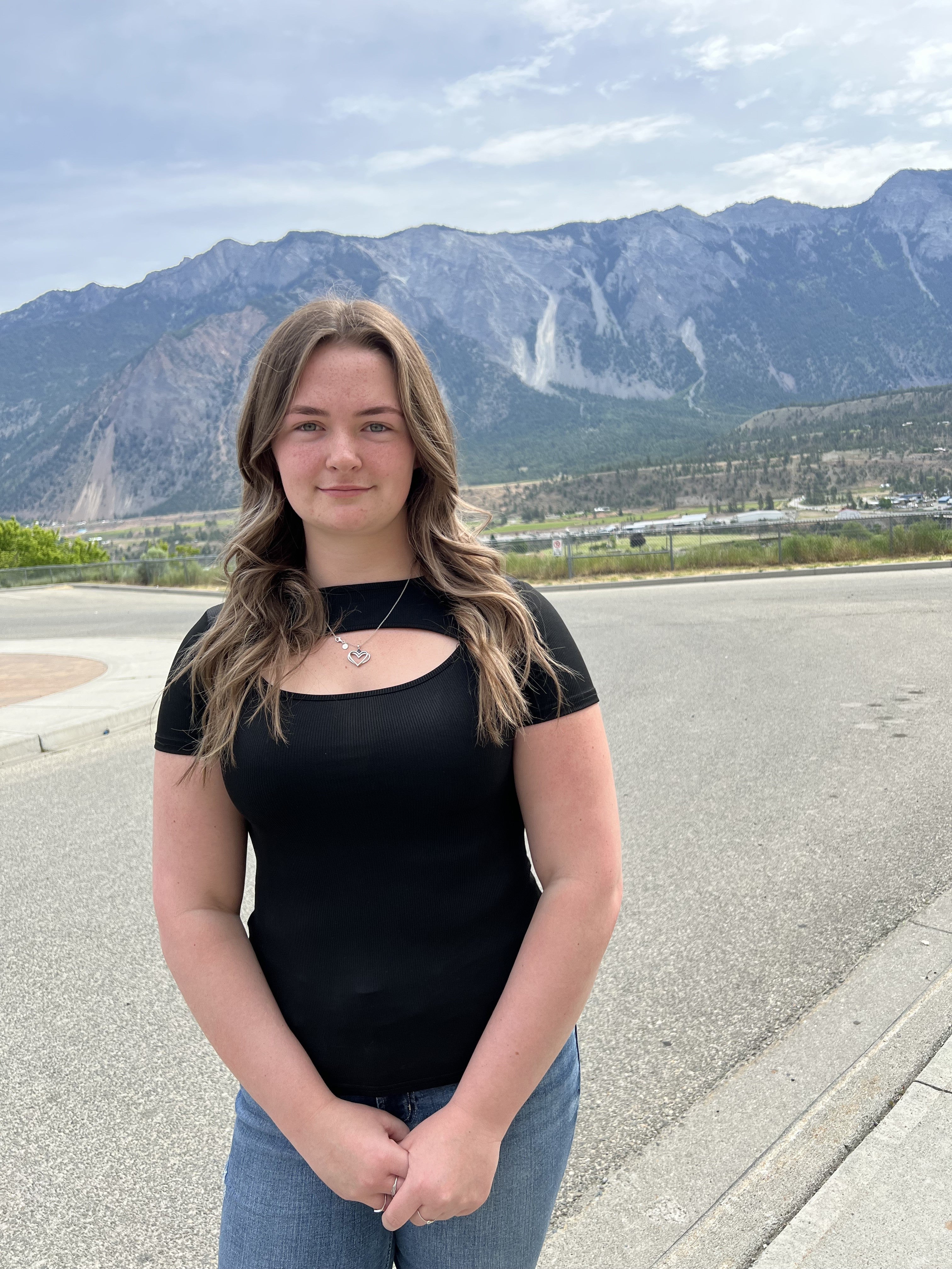 A teenaged girl in a black top stands with her hands together in a parking lot with tall mountains in the background.