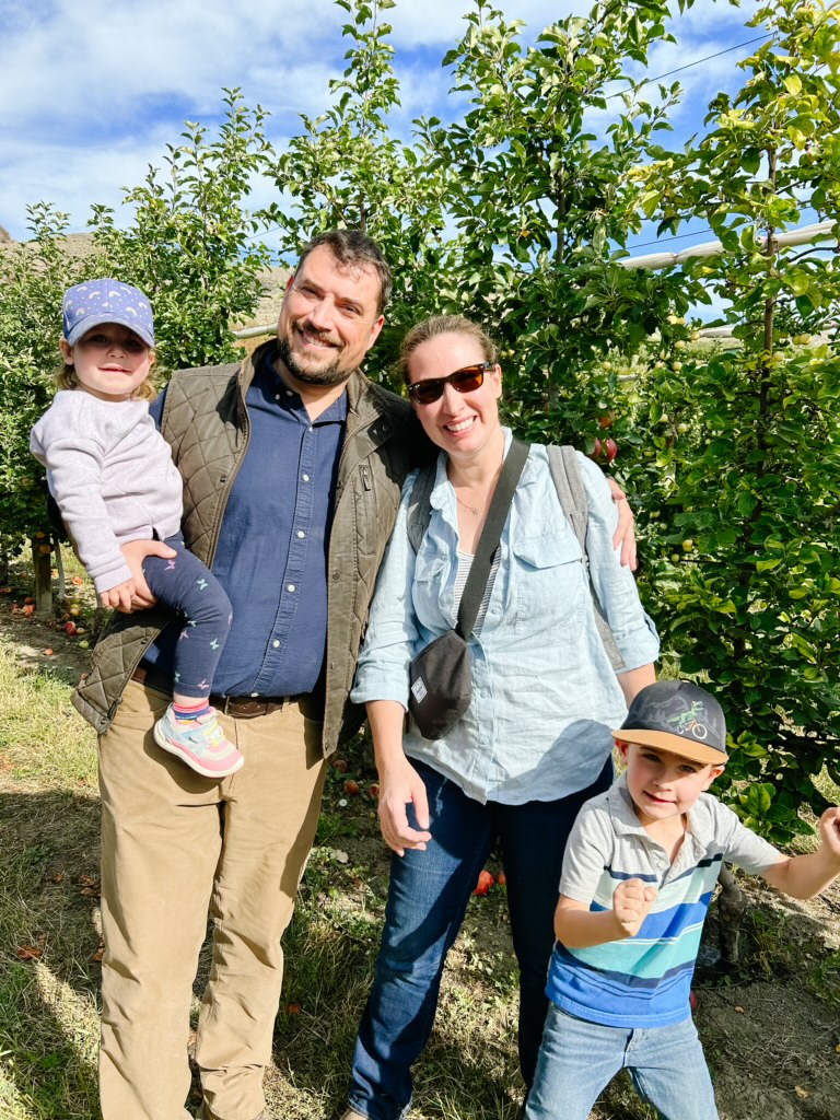 A father, mother, son, and daughter pose in front of fruit trees at an orchard. in Vernon, B.C. The mother has blonde hair pulled back, and wears sunglasses, a light blue chambray shirt, blue jeans, and a black bag. The father wears a blue button-up shirt, khaki pants, and a green vest. The father holds his daughter who wears a purple baseball cap, long-sleeve shirt, navy leggings, and colourful shoes. The son stands in front of the mother, and wears a baseball cap, a striped polo tee, and blue jeans. 