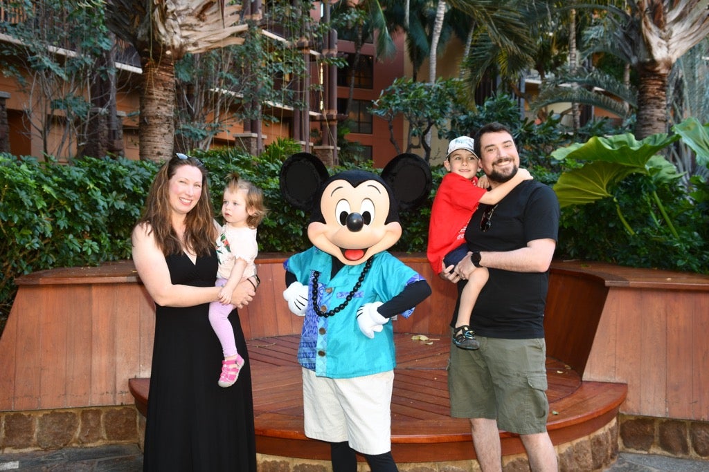 A family of a mother, father, and two young children stands next to Mickey Mouse dressed in tropical attire. The mother, dressed in a black dress, holds a daughter wearing a white t-shirt and purple leggings. The father, dressed in a black tee and khaki green shorts, holds his son wearing a red t-shirt, navy shorts, and a baseball cap. There is tropical greenery and palm trees in the background. 
