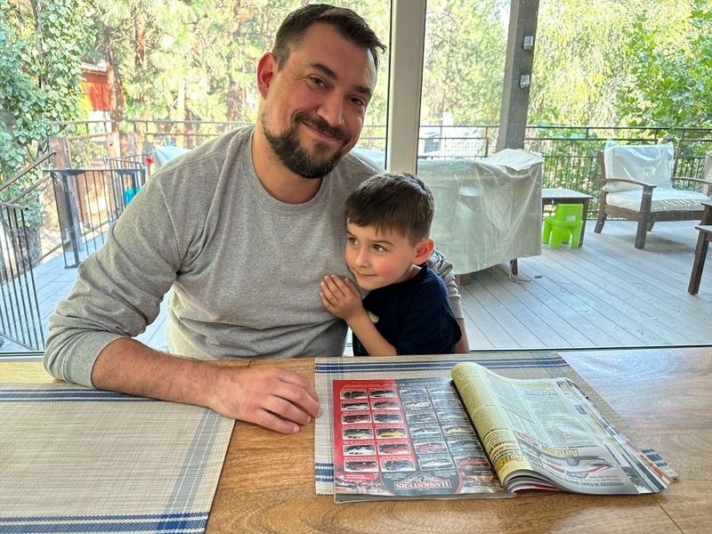 A man with dark hair and facial hair, wearing a grey long-sleeve t-shirt smiles at the camera, while sitting at the table with his arm around a young boy, wearing a navy t-shirt. There is a magazine with many images of cars open on the table in front of them. 