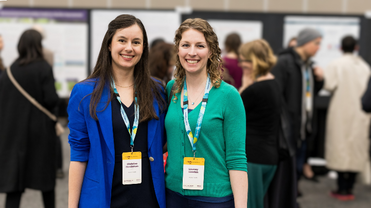 Two people in business attire and wearing lanyards stand together in a conference room.