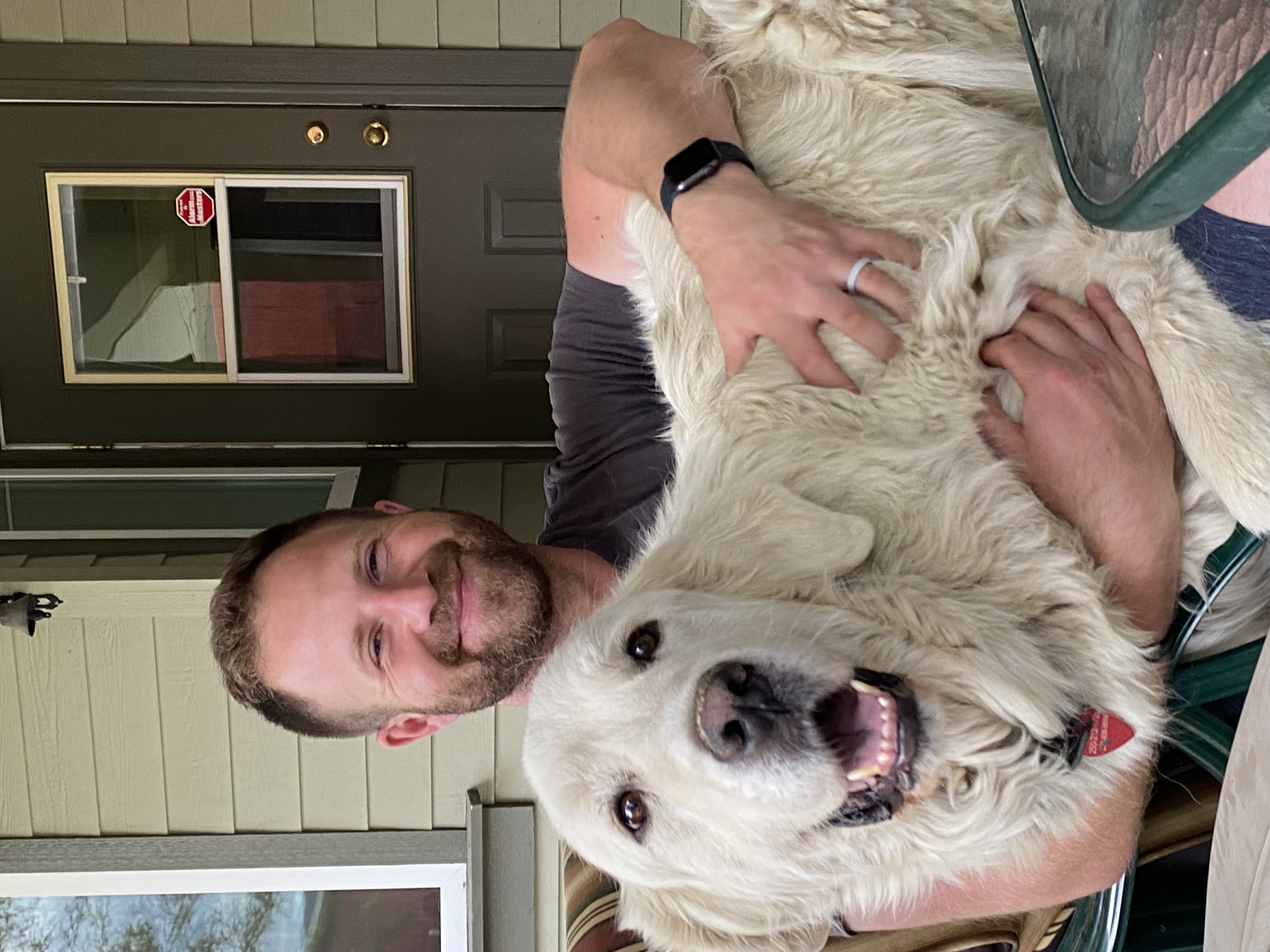 A person with a beard sits hugging a big white furry dog on his lap in front of a door.