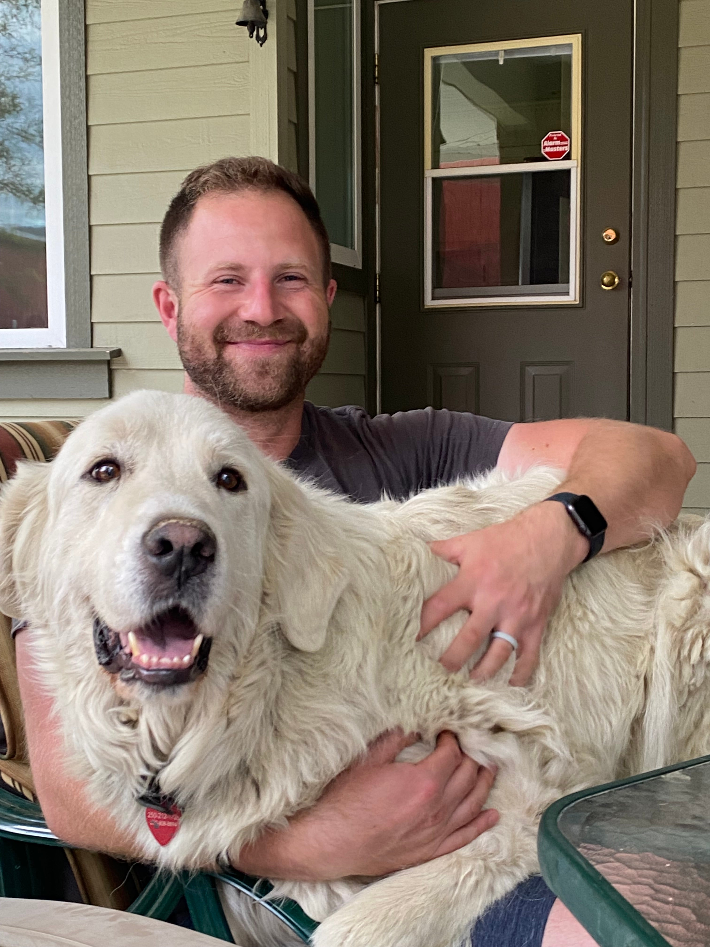 A person with a beard sits hugging a big white furry dog on his lap in front of a door.