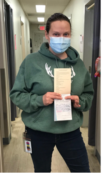 A person wearing a mask and a green hoodie standing in a hallway holds a prescription pad