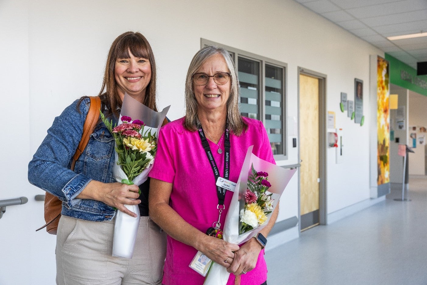 Two people holding bouquets of flowers, one in a jean jacket, one in a bright pink top, stand in a hallway.
