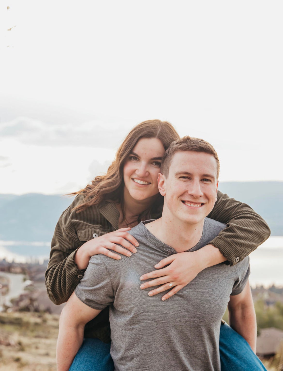 A woman with long brown hair in a brown corduroy shirt piggy backs on a man in a grey t-shirt