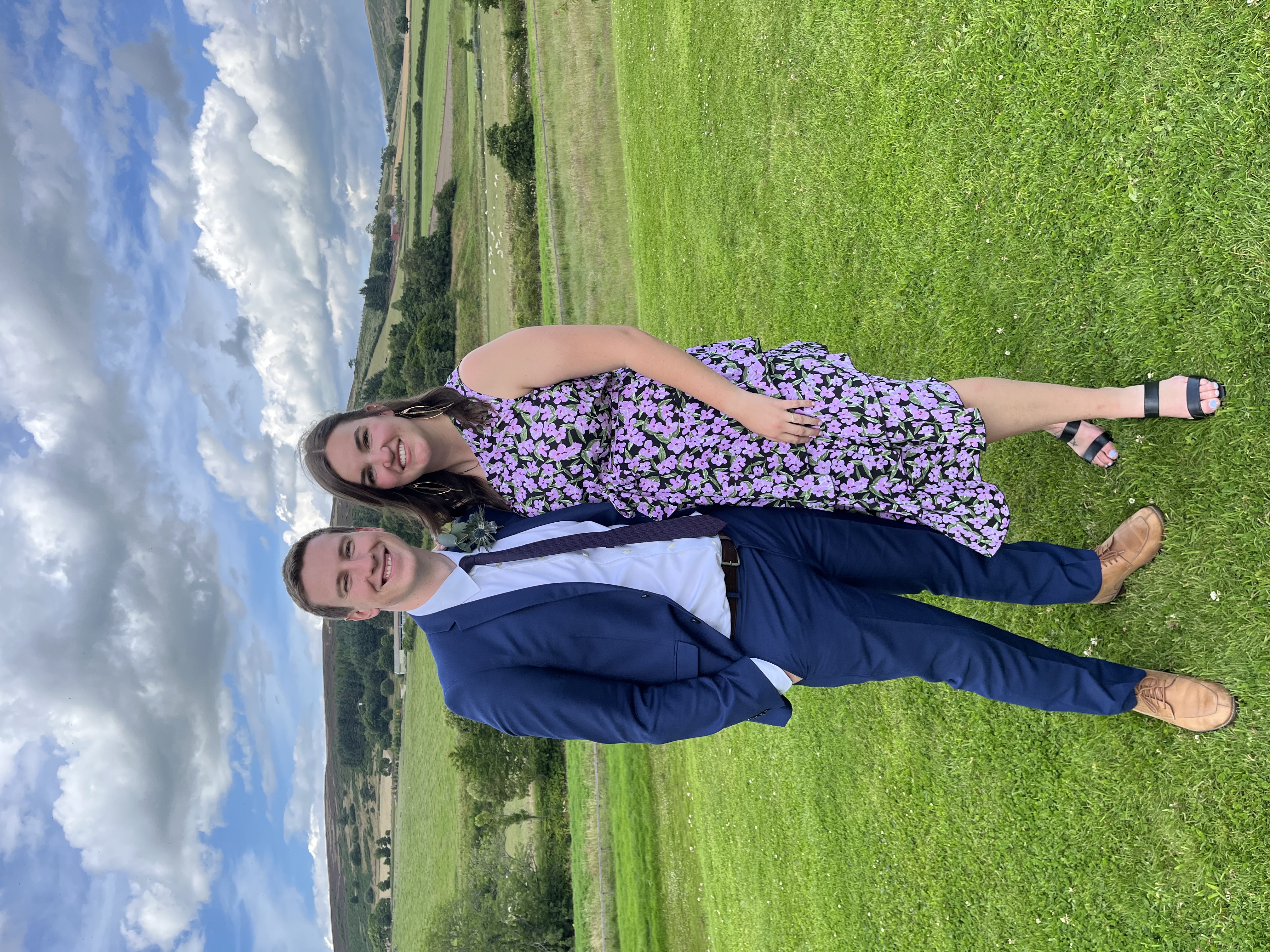 Two people pose in formal attire in a grassy fields surrounded by hills and blue and cloudy sky