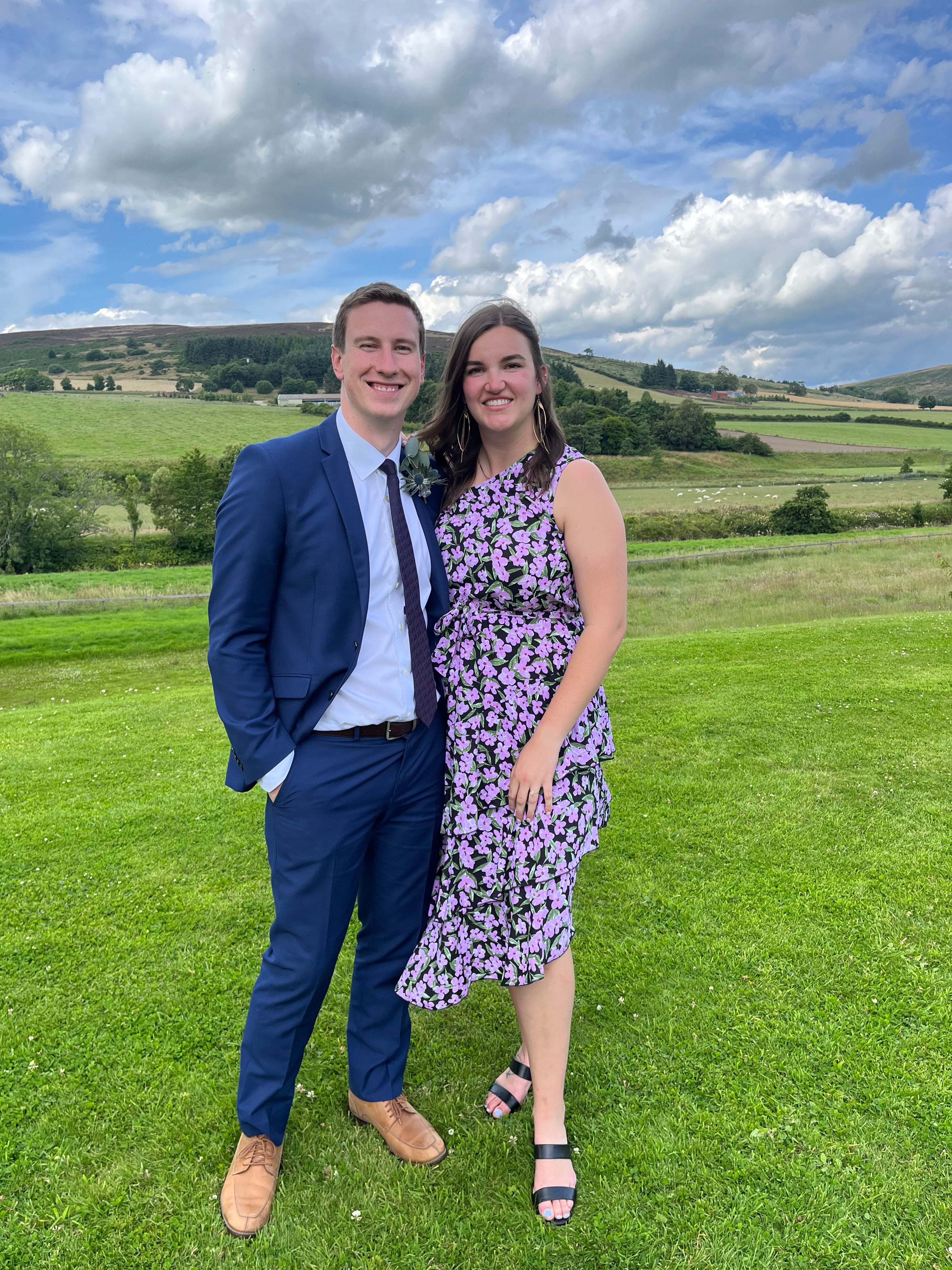 Two people pose in formal attire in a grassy fields surrounded by hills and blue and cloudy sky