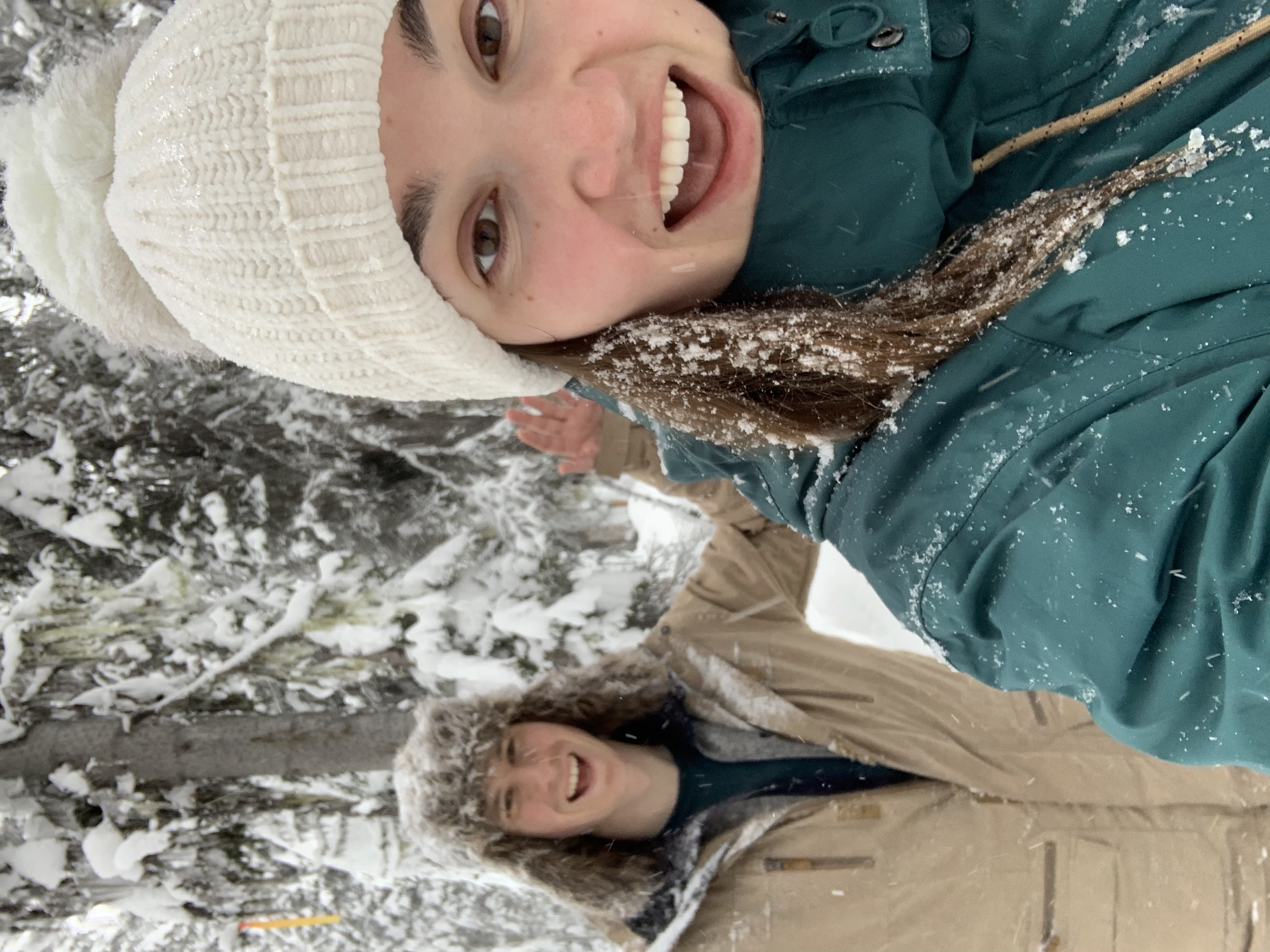Two people wearing winter hats and jackets pose for a selfie photo in a snowy forest.