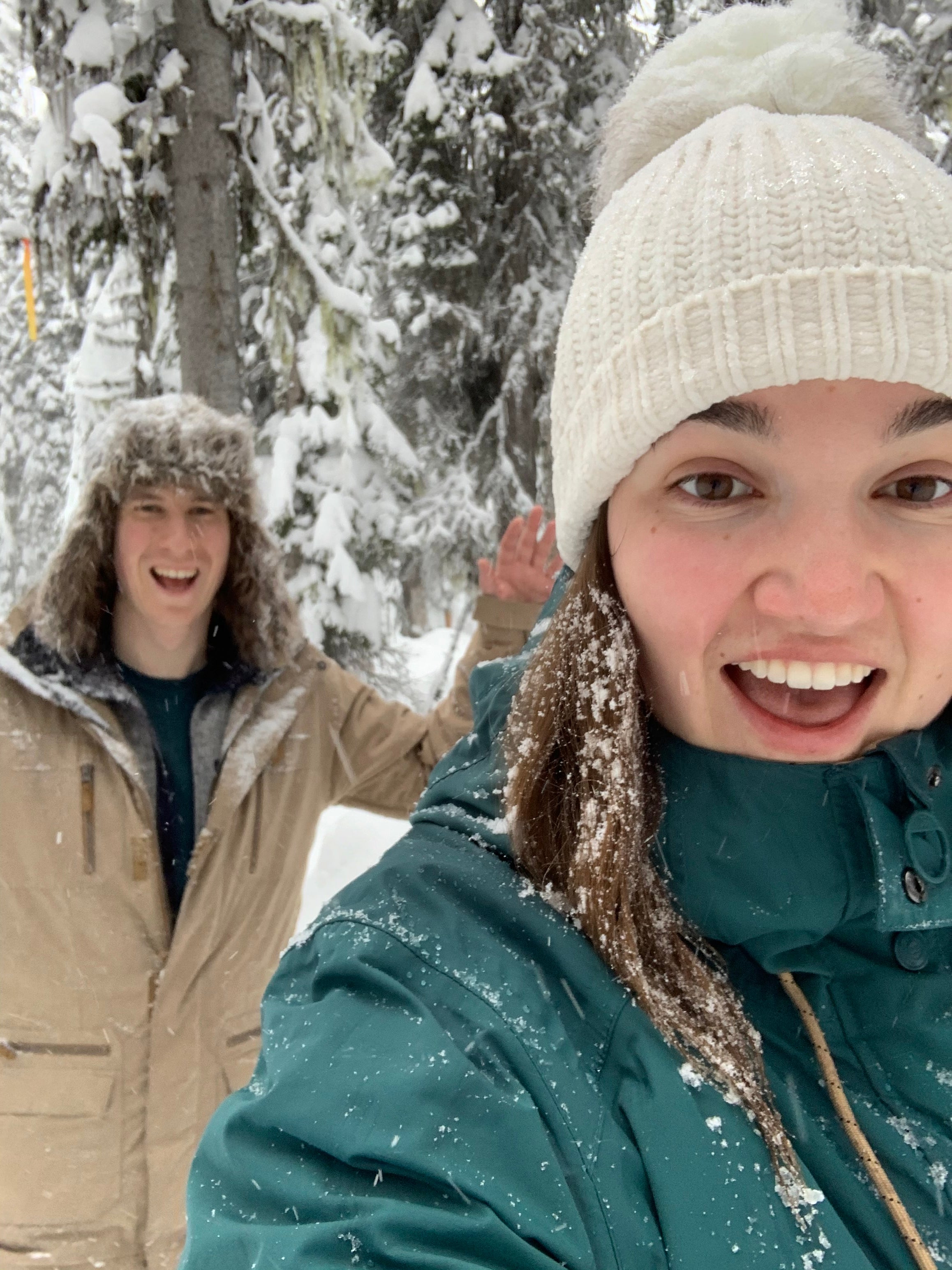 Two people wearing winter hats and jackets pose for a selfie photo in a snowy forest.