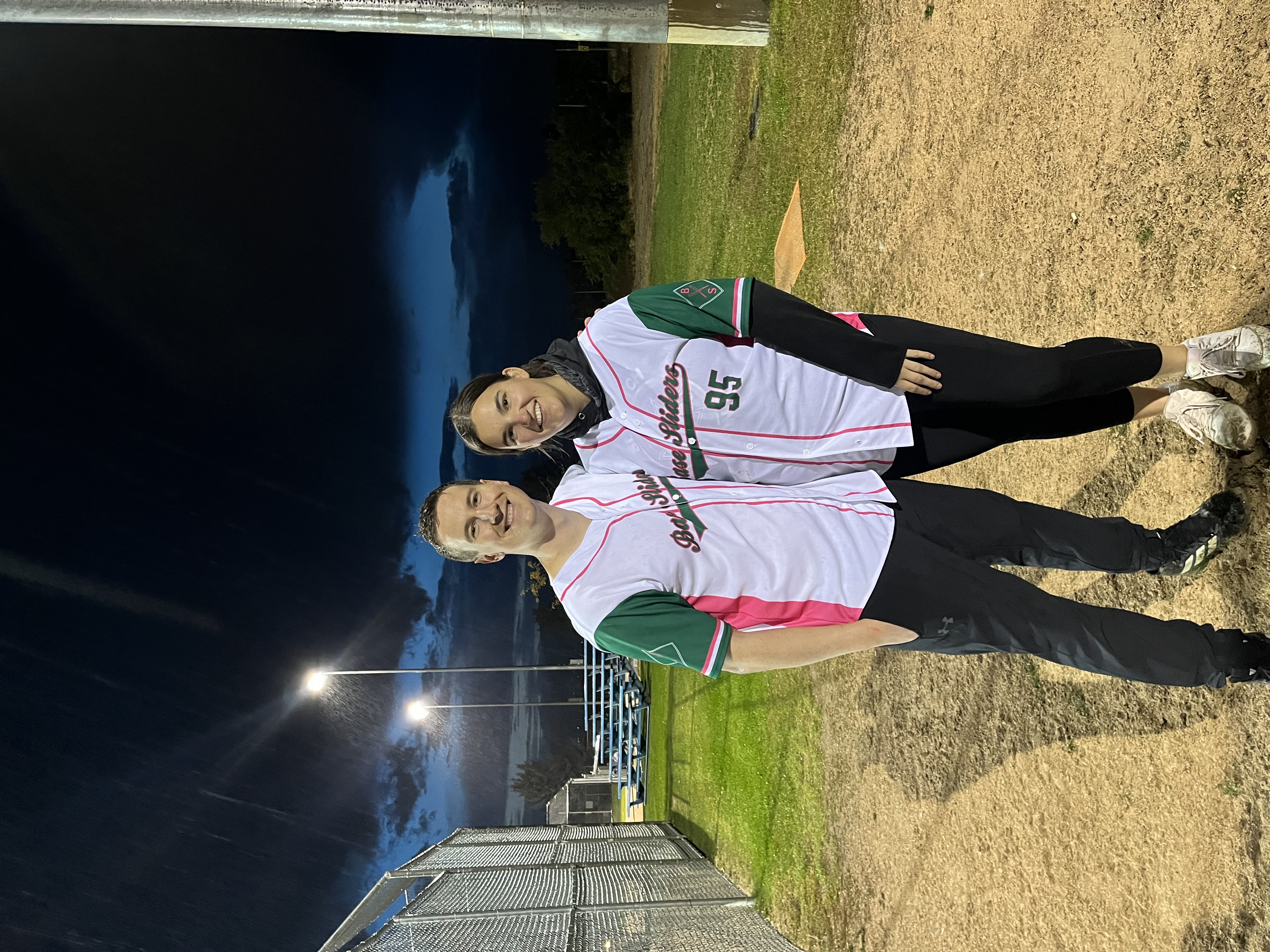 Two people stand together wearing softball jerseys at a ballpark diamond in the evening