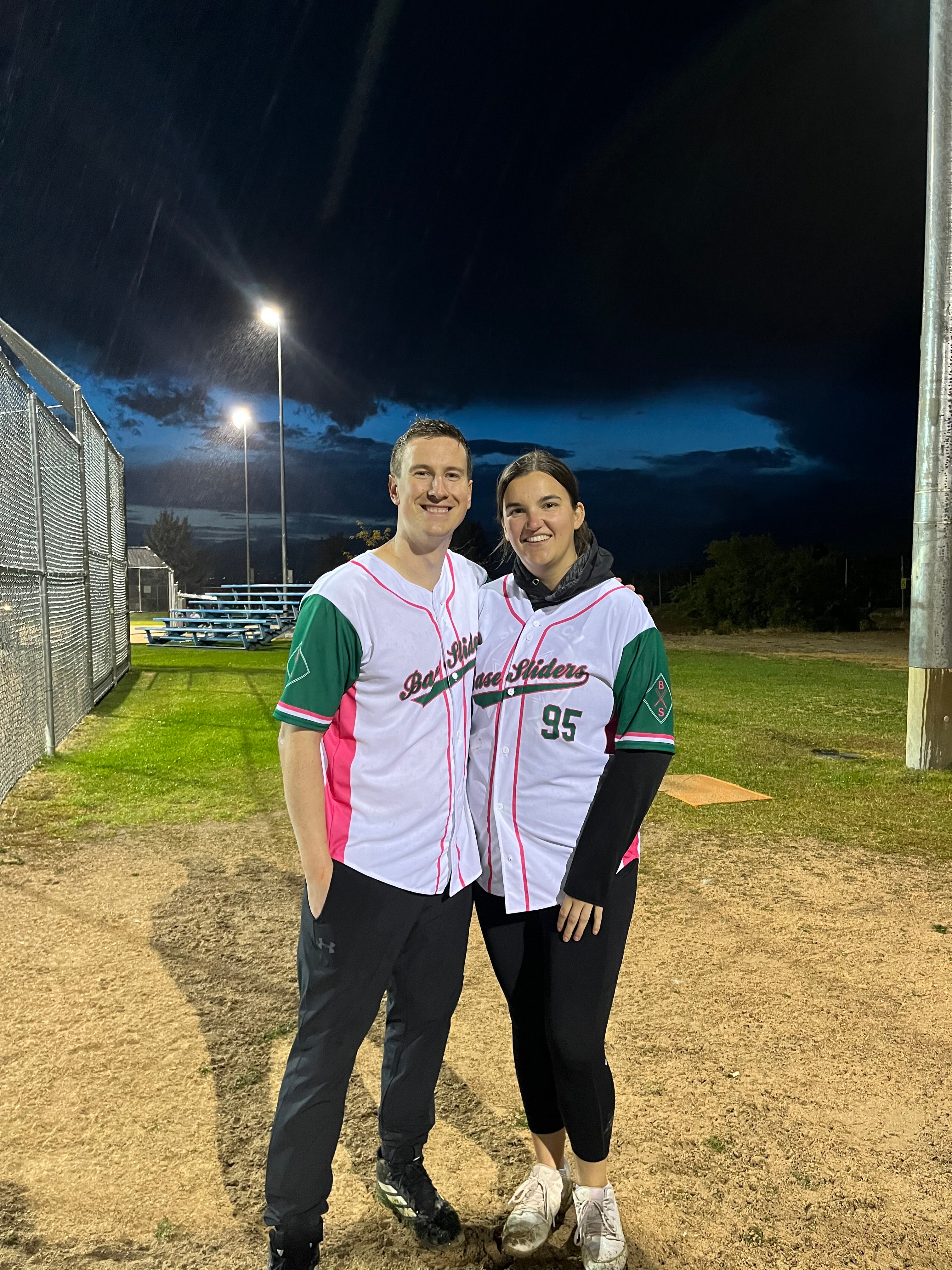 Two people stand together wearing softball jerseys at a ballpark diamond in the evening