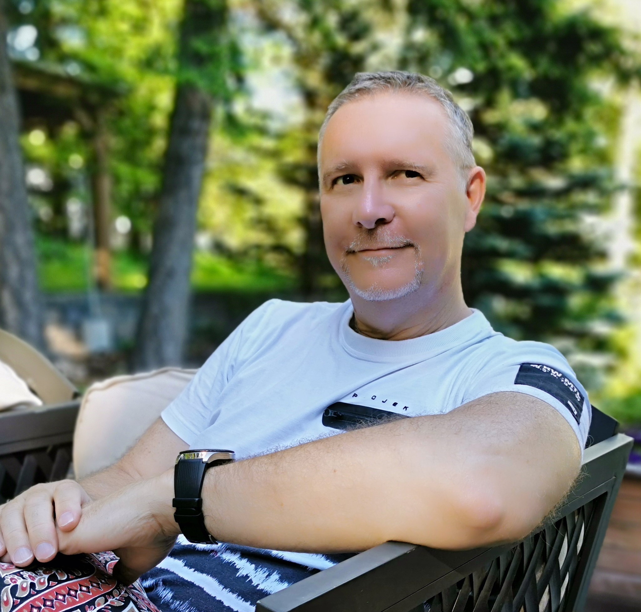 Man with grey hair sits outdoors on a chair and smiles.