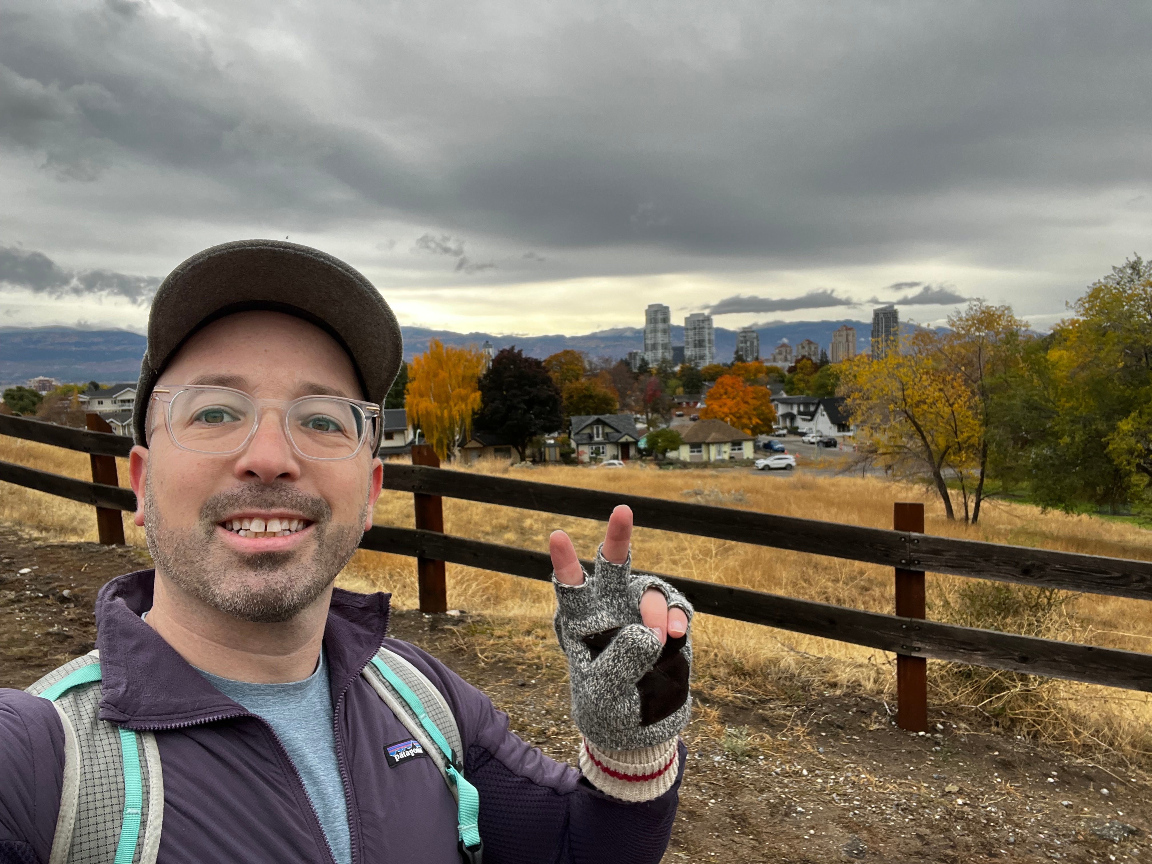 A person with a ballcap, glasses, jacket and knapsack gives a peace sign while walking on a trail on a cloudy day.