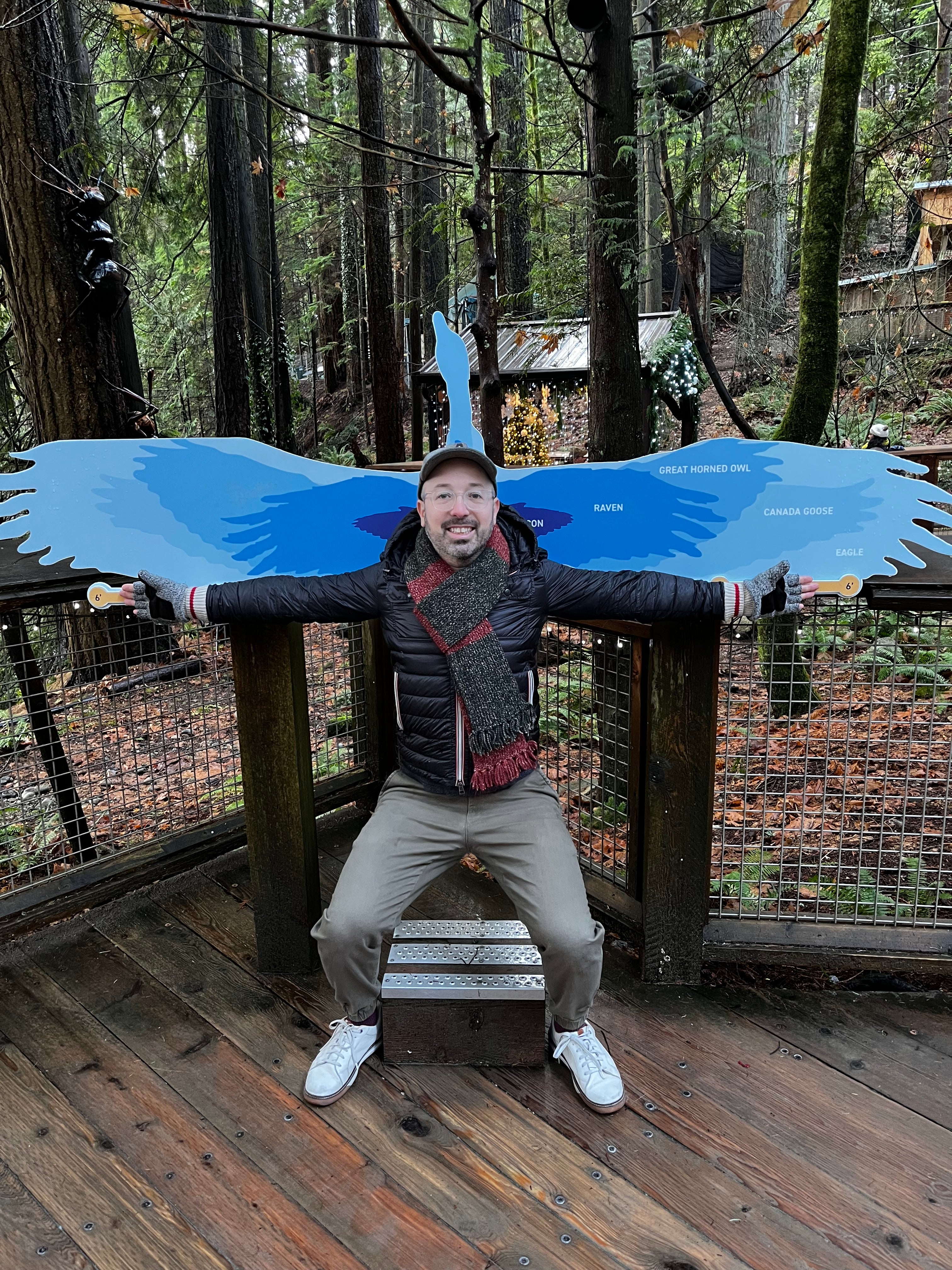 A person sits on a bench in a forest. Their arms are spread out in front of the blue wings with markings of the wingspan of different birds.