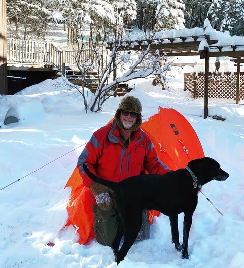 A man in a winter hat and red jacket sits in front of an orange tent with his dog.