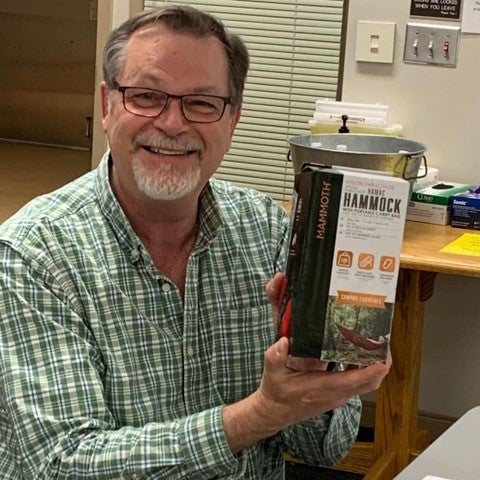 A bearded man wearing glasses in a green checkered shirt holds a hammock in a box.