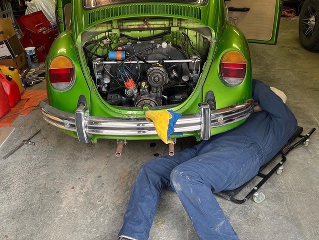 A person in blue overalls lies on a dolly under a green Volkswagen Beetle with its hood open.